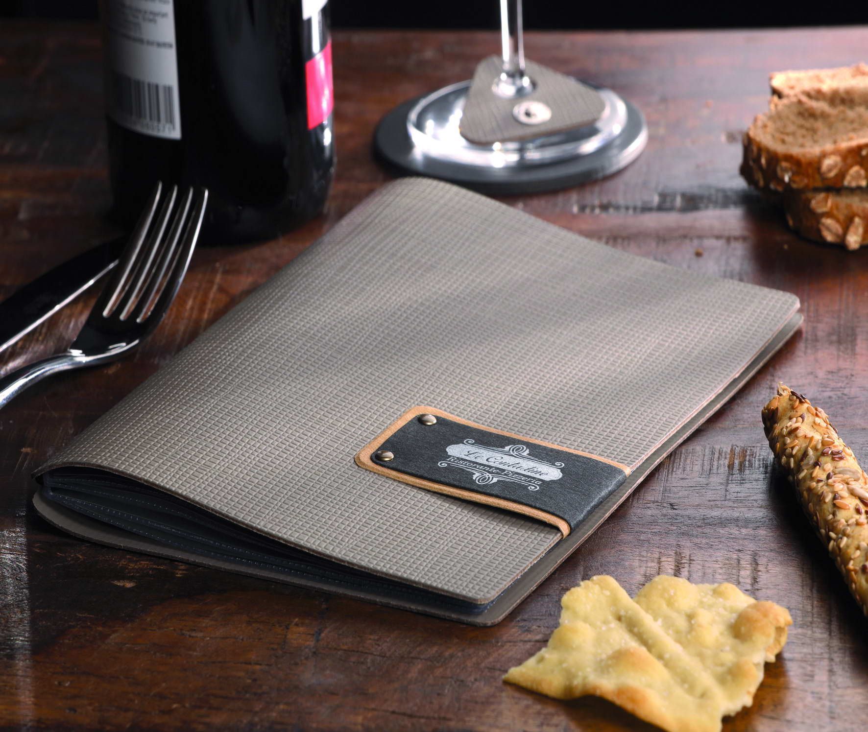 Un menu relié en cuir repose sur une table en bois, à côté d'une bouteille de vin, d'un verre, de pain et de couverts.