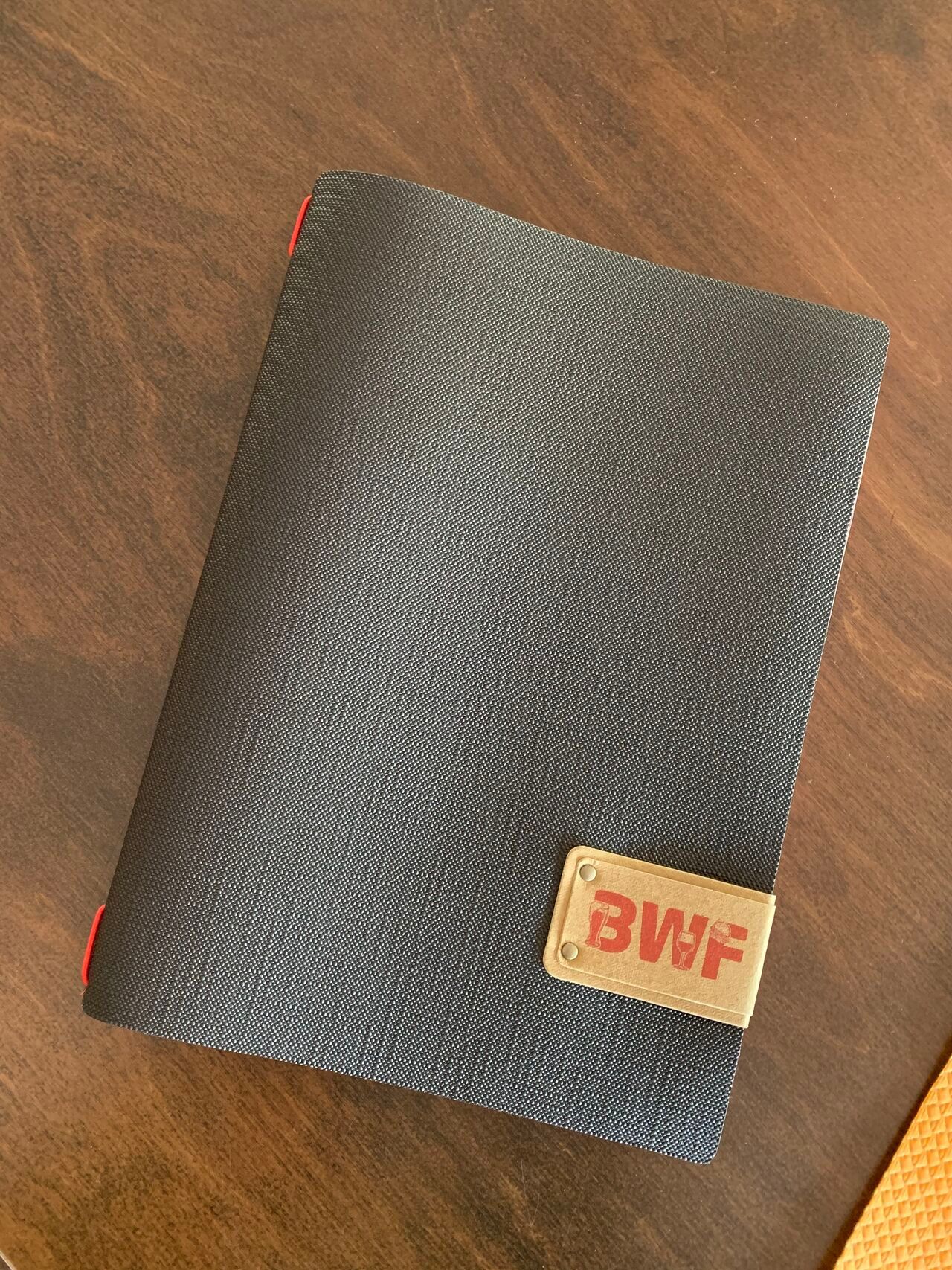 Portefeuille gris foncé avec bordure rouge et logo « BWF » sur une surface en bois.