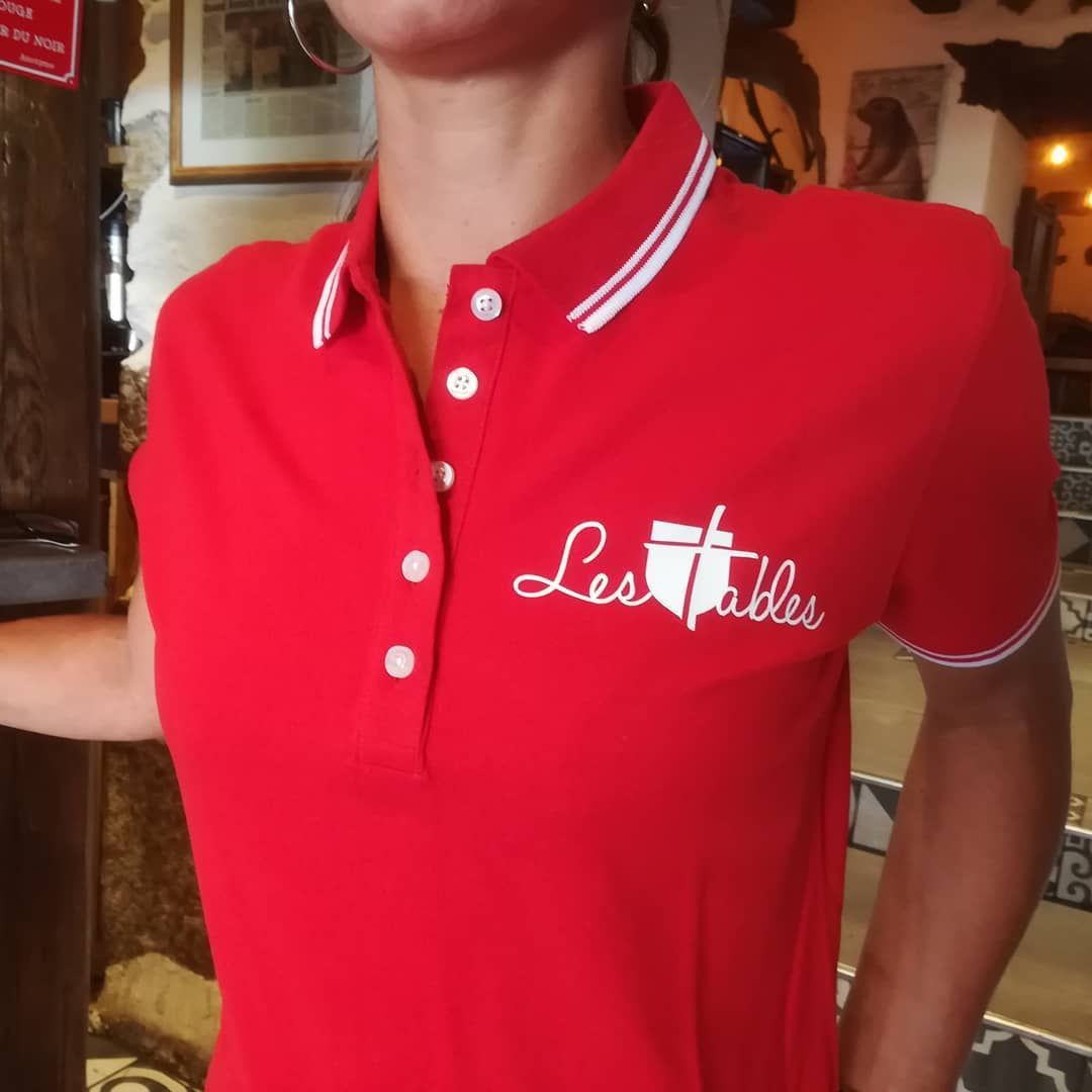 Femme portant un polo rouge avec le logo « Les Tables », une bordure blanche et des boutons.
