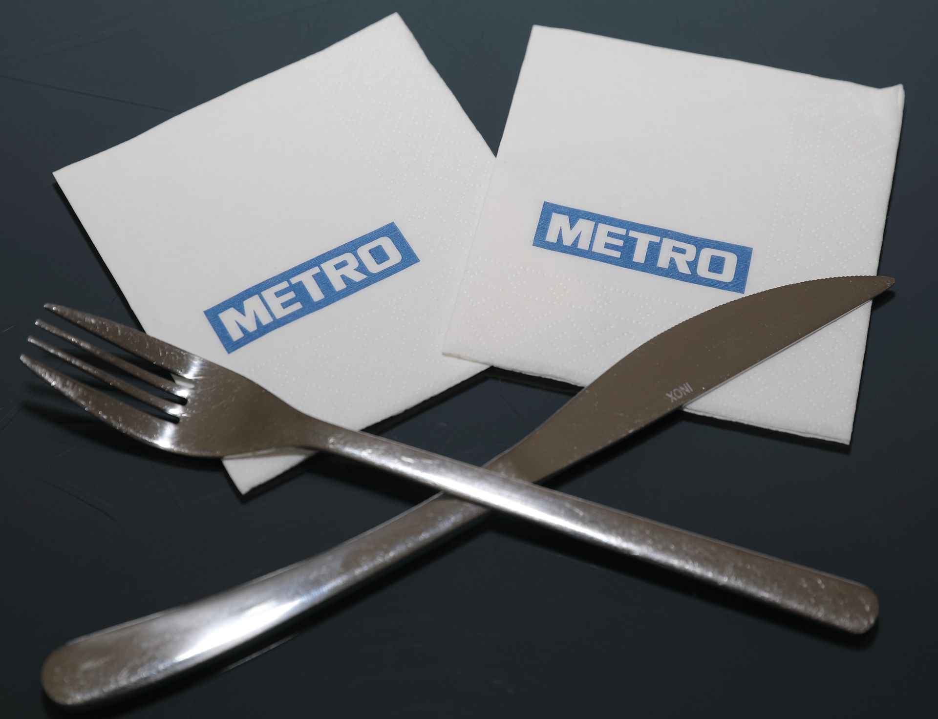 Fourchette et couteau croisés sur une surface sombre, avec deux serviettes blanches portant le logo « METRO ».