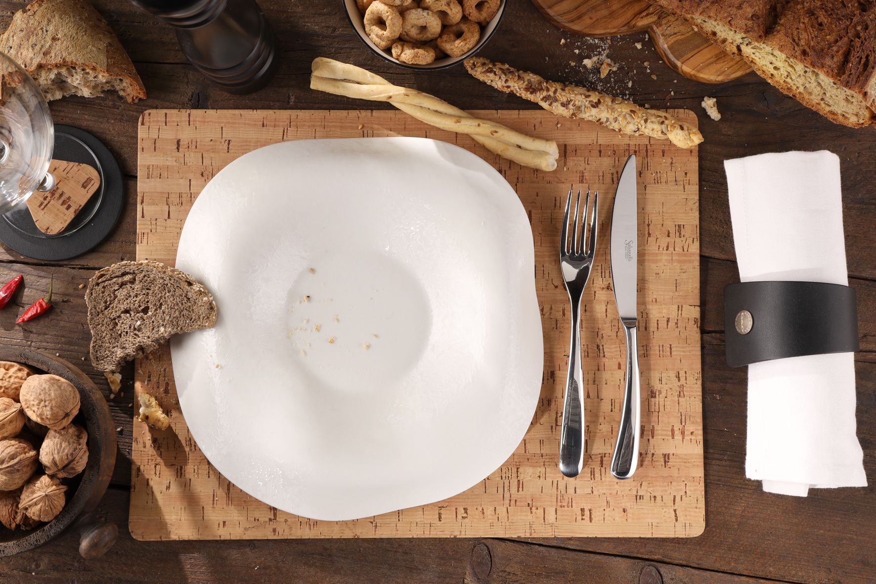 Assiette blanche sur tableau en liège avec couverts, rond de serviette, pain, noix et collations.