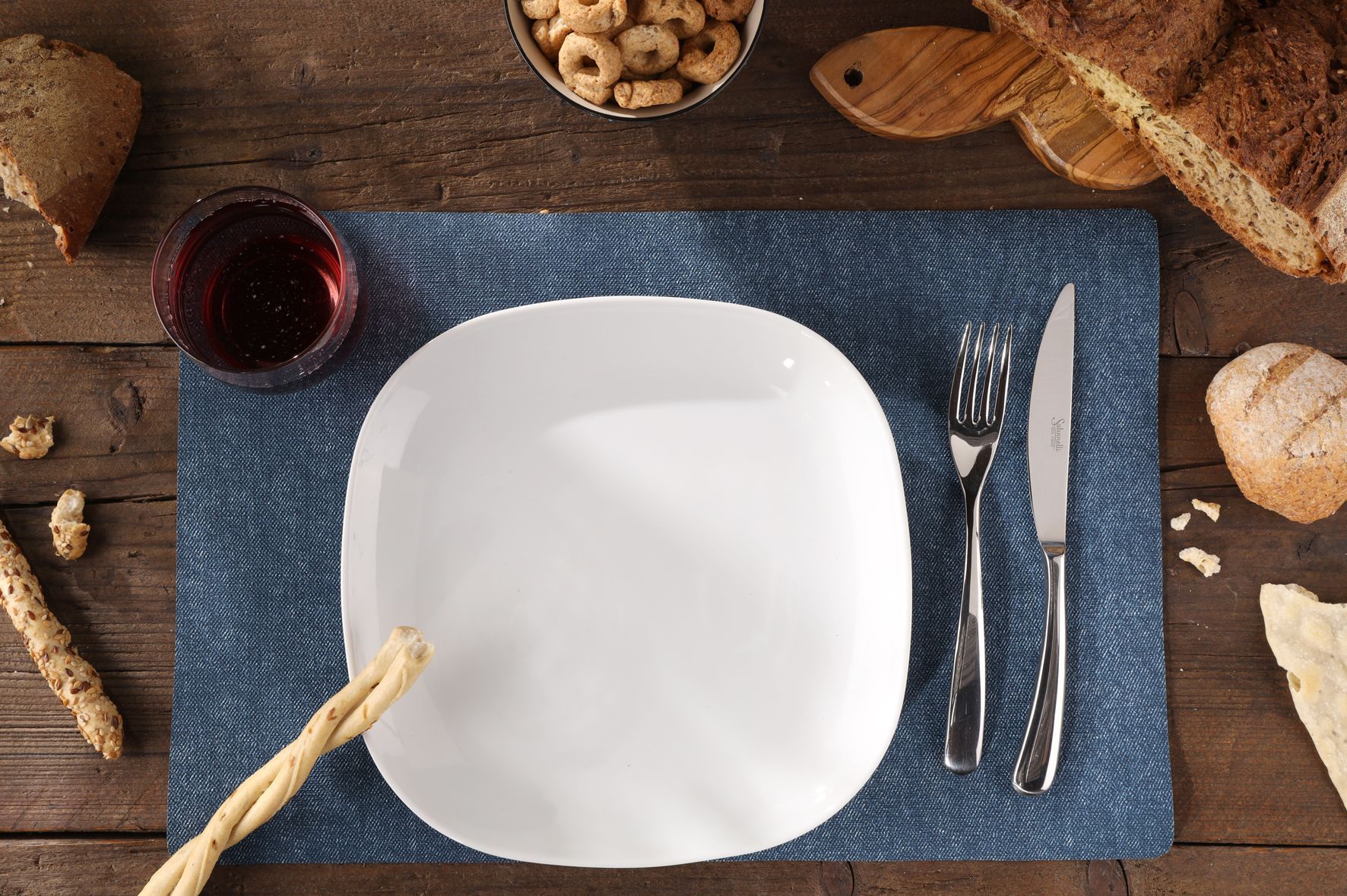 Table dressée avec assiette, couverts, vin, pain et collations sur une surface en bois.