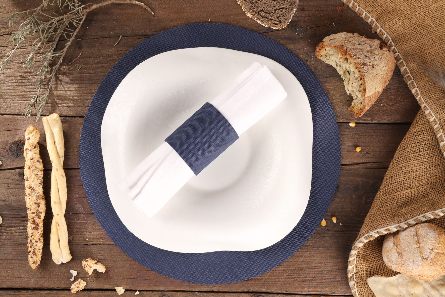 Assiette blanche avec serviette, pain et gressins sur un set de table bleu et une table en bois.