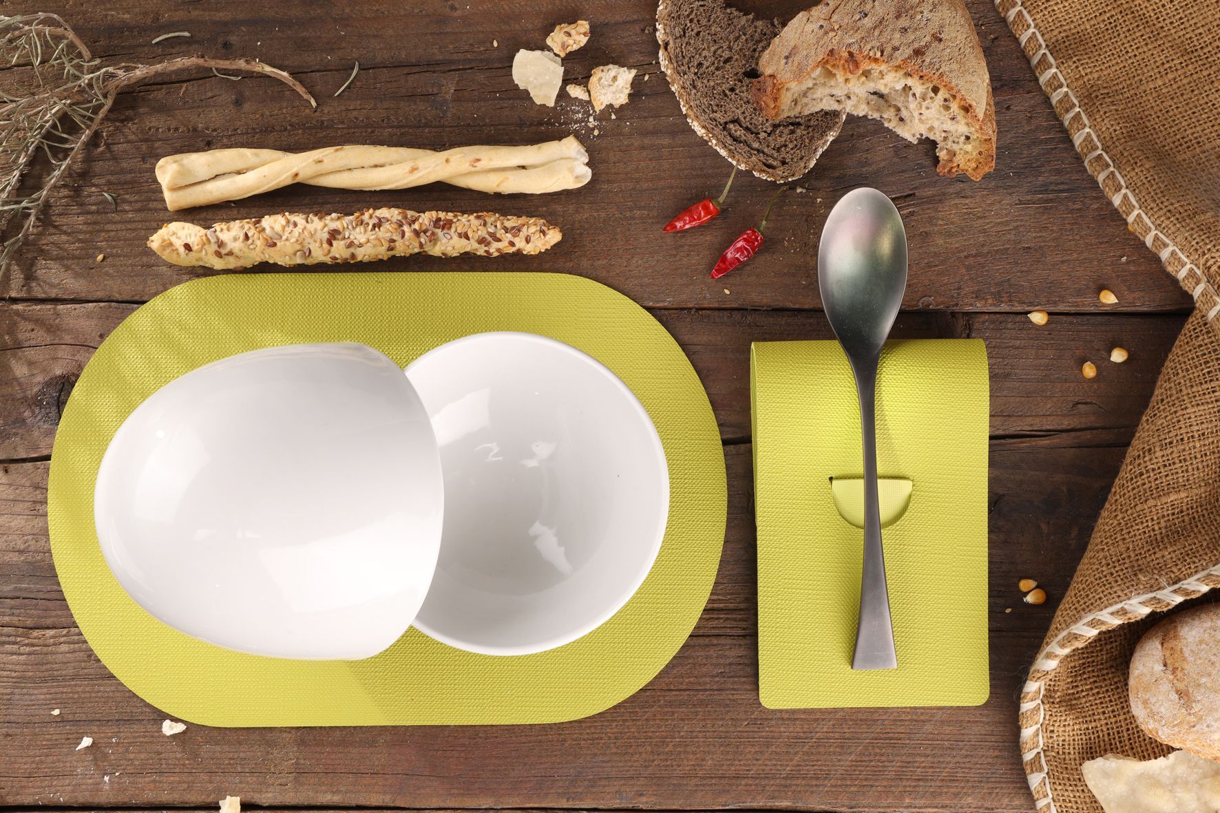 Deux bols blancs sur un set de table jaune, avec du pain, des gressins et une cuillère sur une table en bois.