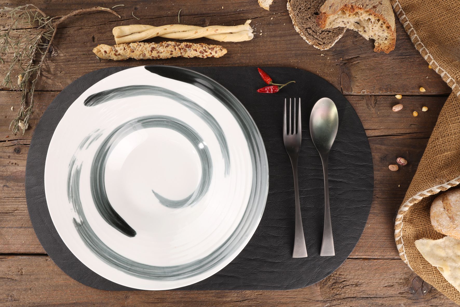 Bol blanc avec motif tourbillon gris, couverts et pain sur une table en bois.