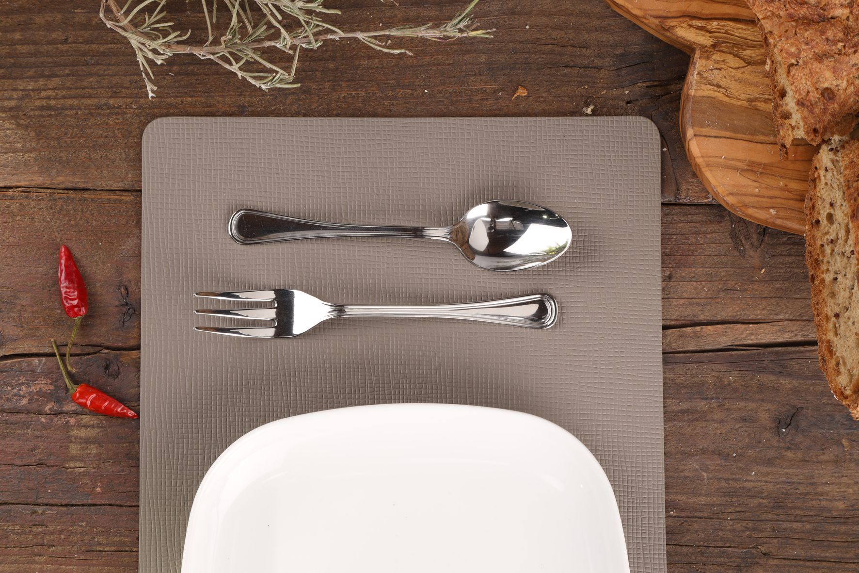 De l'argenterie sur un set de table gris avec une assiette blanche, des poivrons rouges et du pain sur une surface en bois.