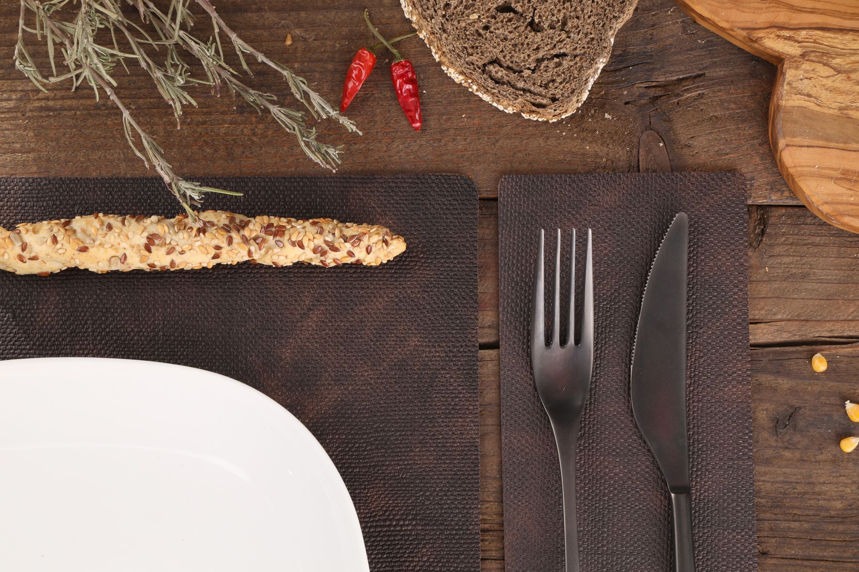 Une table rustique avec une assiette, un gressin, du pain, une fourchette, un couteau et des herbes.