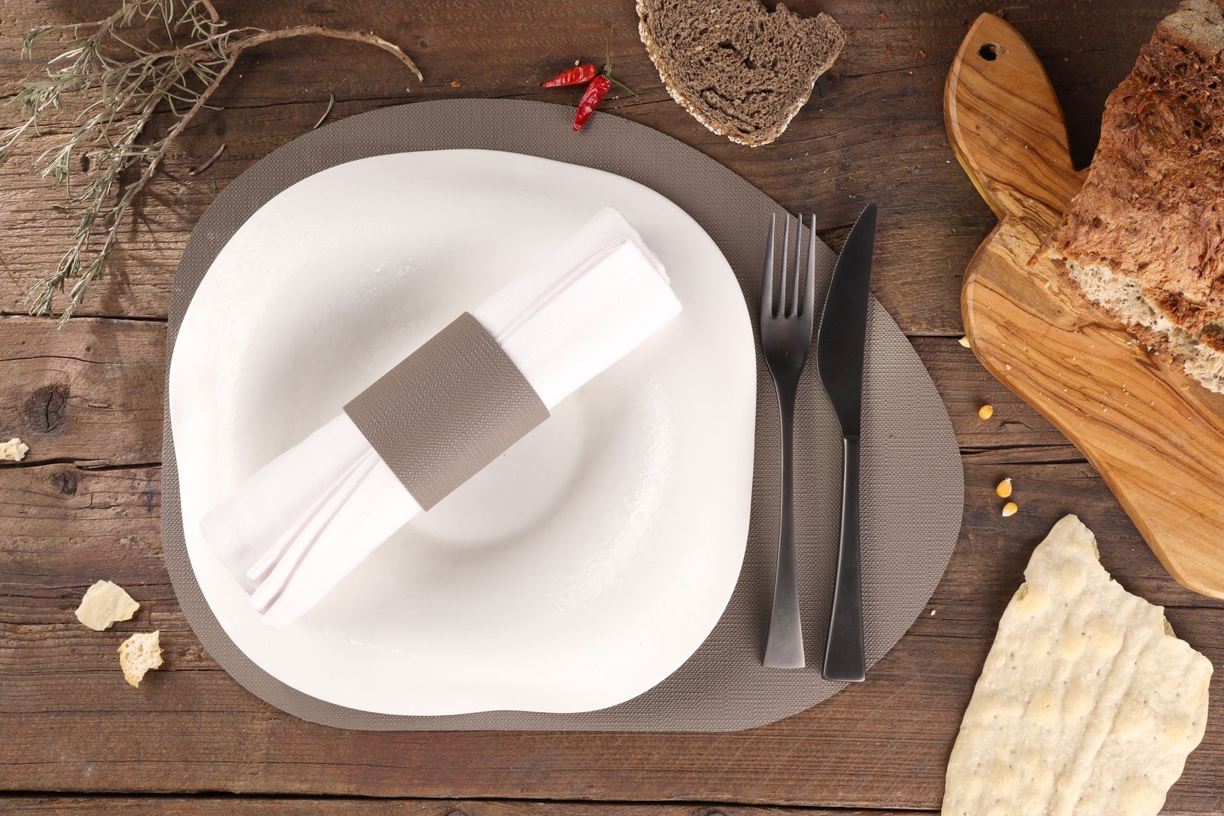 Couvert avec assiette blanche, serviette, ustensiles, pain et piment sur une table en bois.