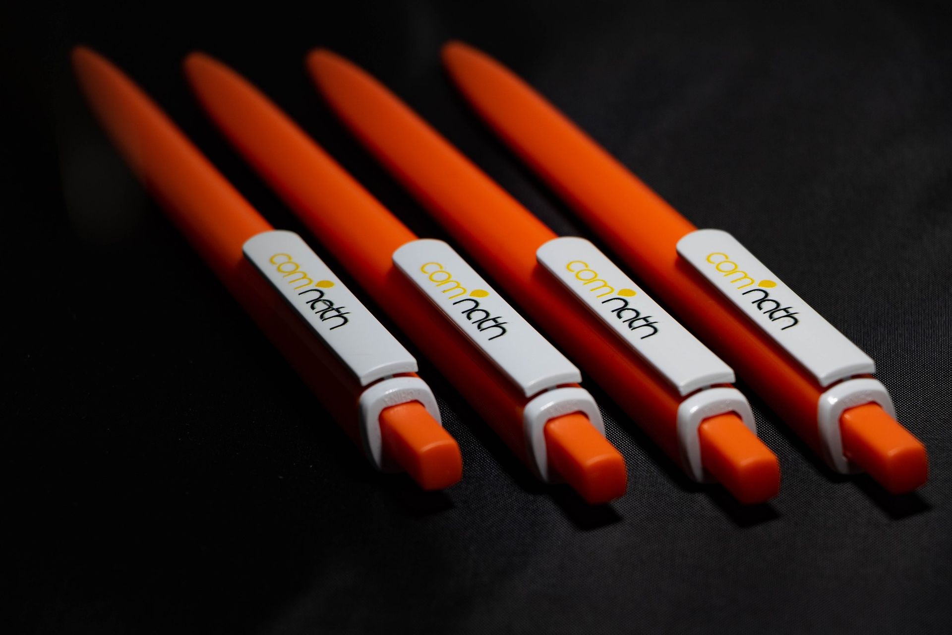 Quatre stylos orange disposés en diagonale sur une surface sombre, chacun avec une étiquette et un logo blancs.