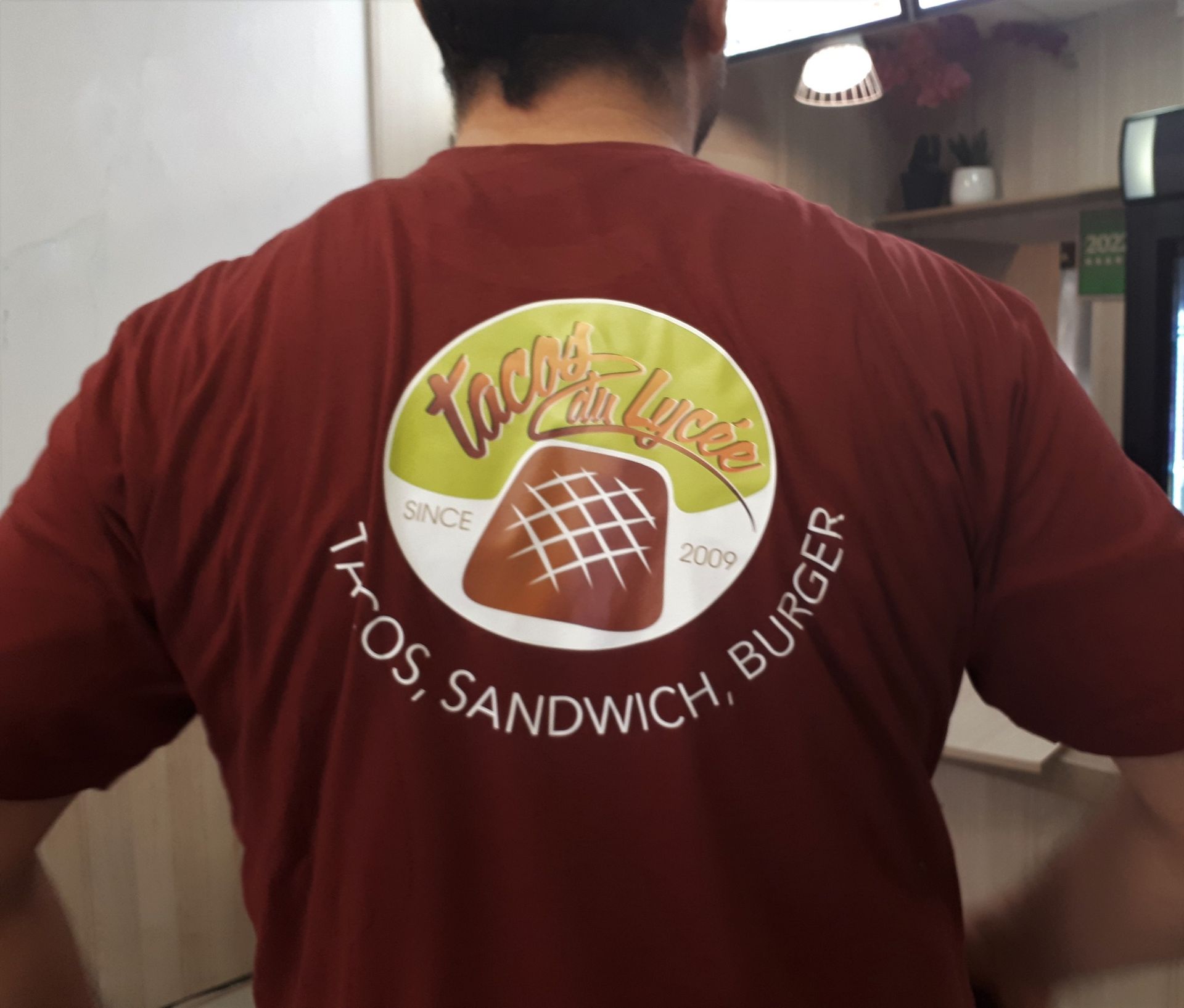 Personne portant un t-shirt marron avec un logo « Tacos Can Lycee » dans le dos.