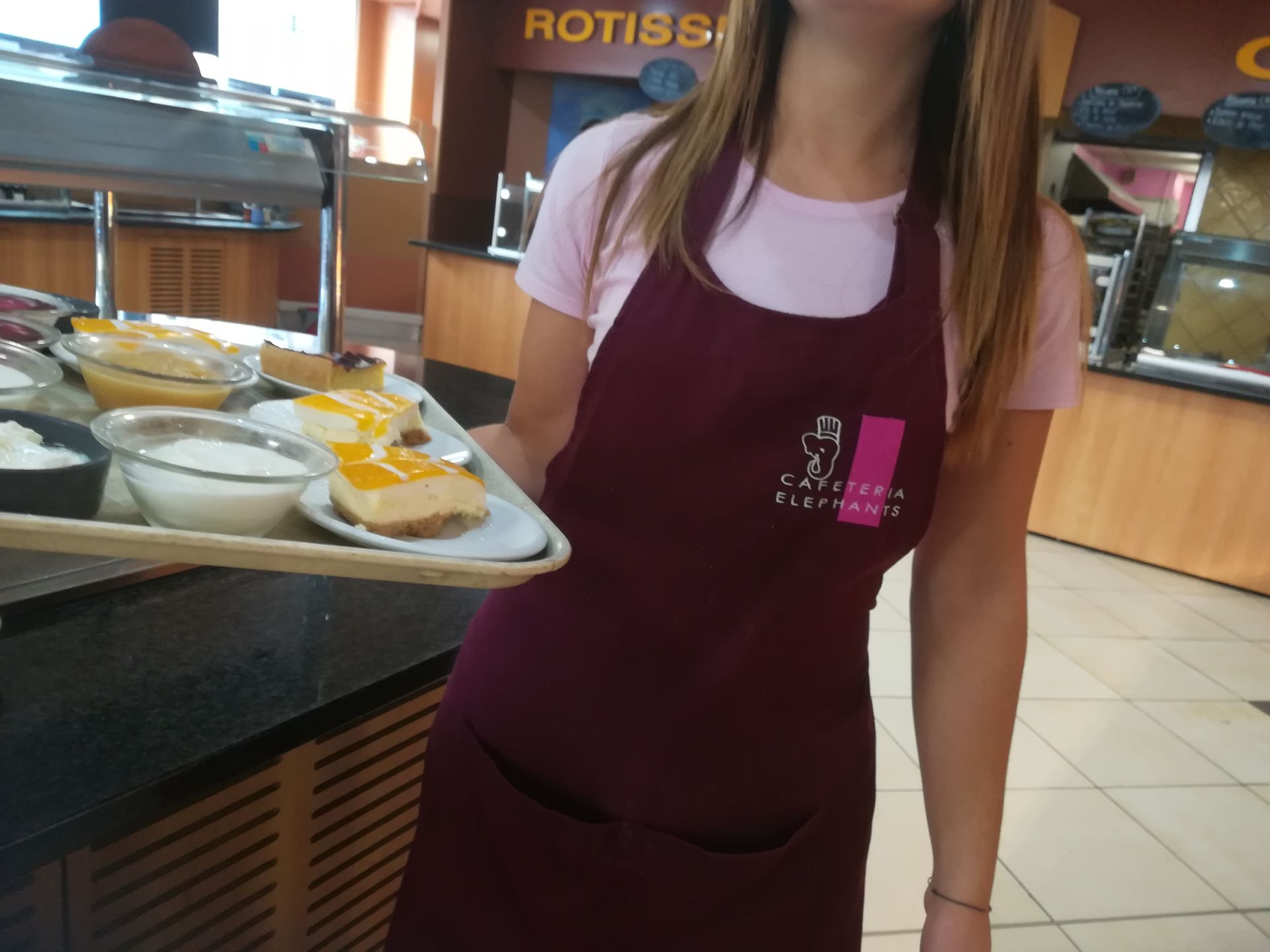 Femme en tablier marron tenant un plateau de desserts dans un restaurant.
