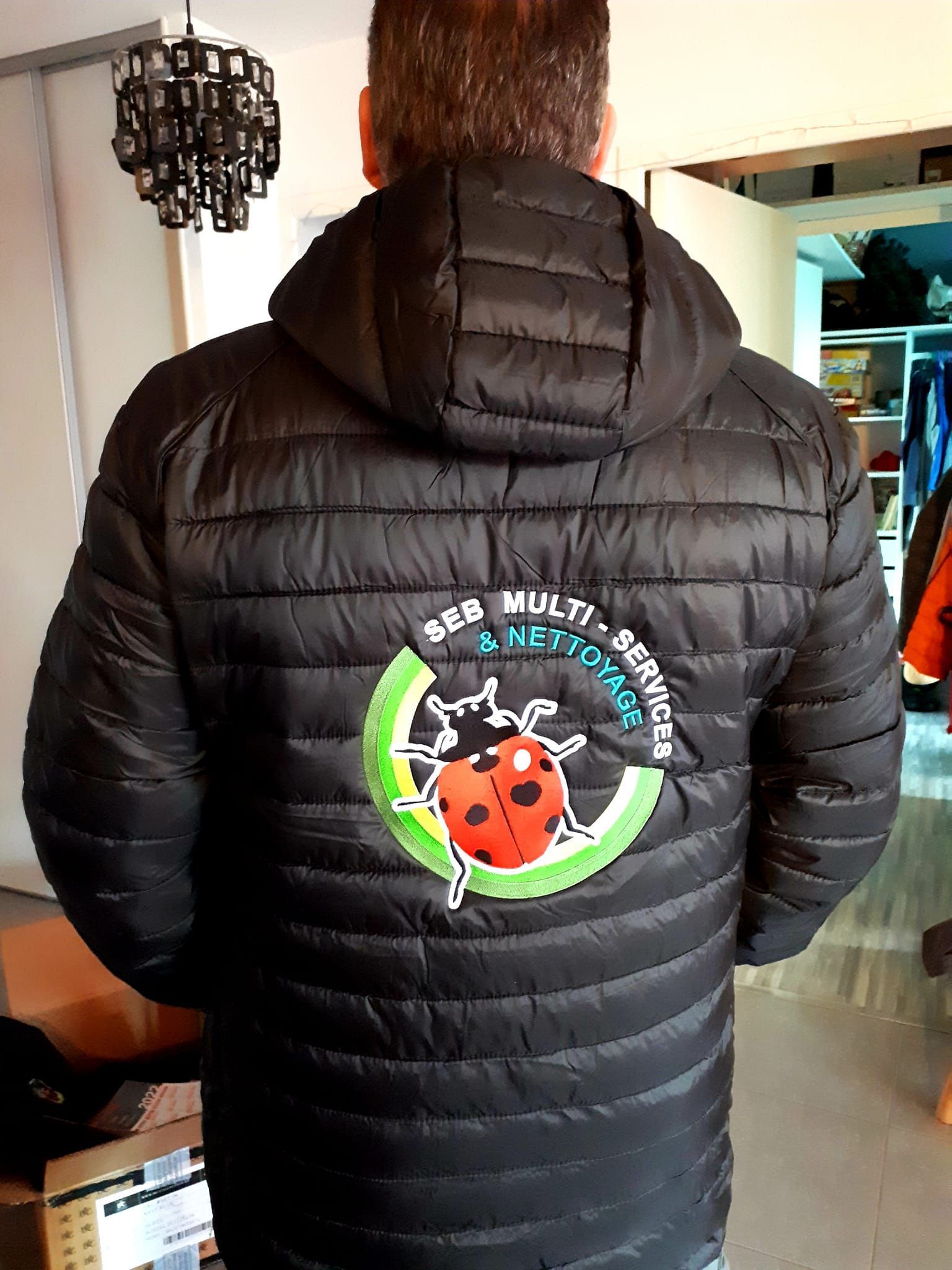 Personne portant une doudoune noire avec un logo de coccinelle dans le dos, dans un cadre domestique.