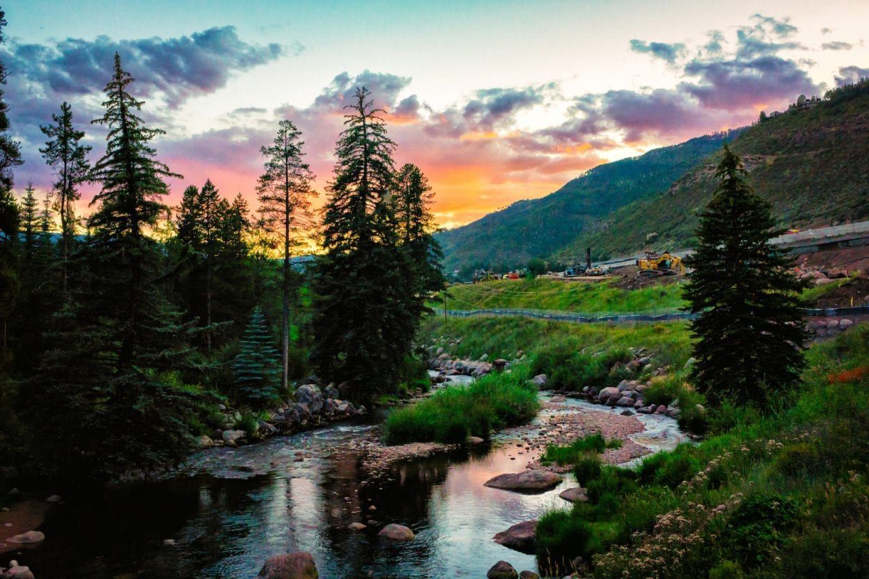 Mountain stream at sunset with pine trees, rocky banks, and a colorful sky over distant hills