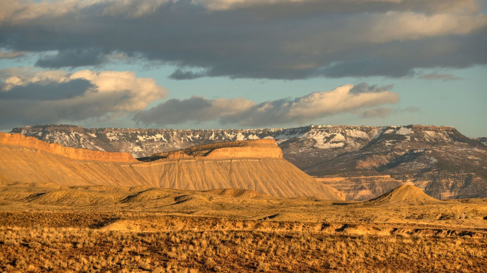 Sunlit desert mesas under dramatic clouds, with layered cliffs and golden scrubland in the foreground