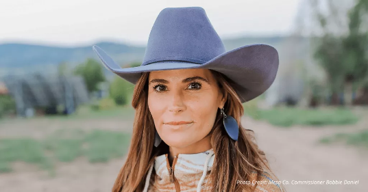Woman in a lavender cowboy hat outdoors, with a blurred rural landscape behind her.