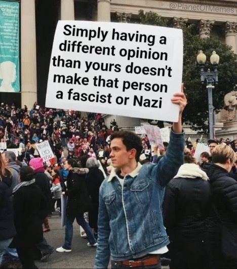 Protester in denim jacket holding a sign at a crowded rally: “Simply having a different opinion...”