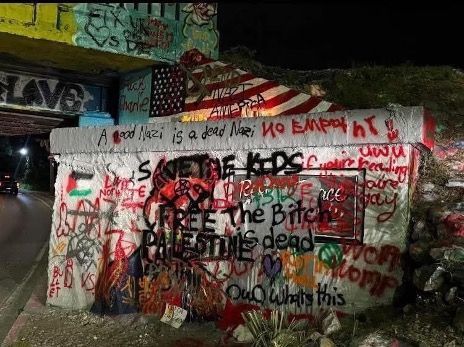 Graffiti-covered concrete wall at night with red, black, and white protest-style writing and posters