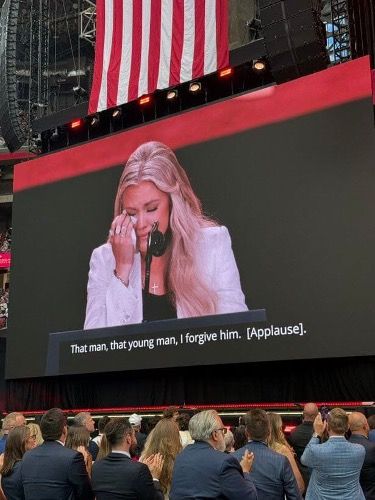 Large stadium screen shows a blonde woman speaking at a microphone, with crowd below and a U.S. flag overhead.