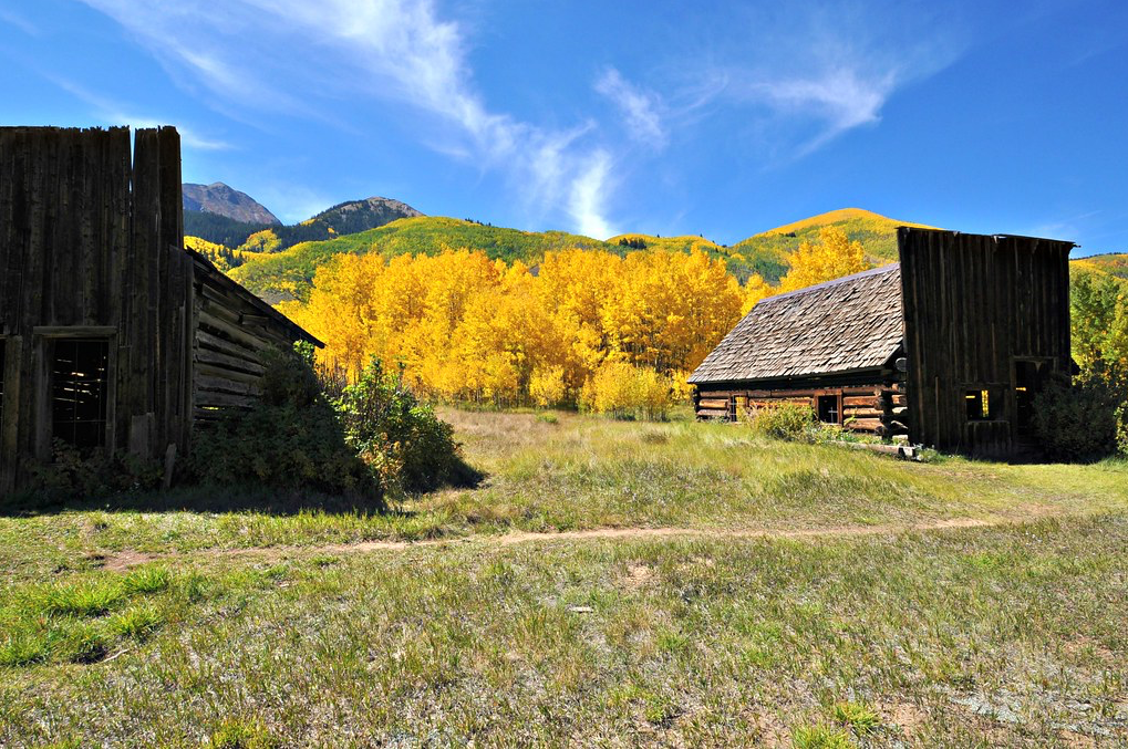 Rustic barns framing a golden autumn forest under a bright blue sky with mountains in the background