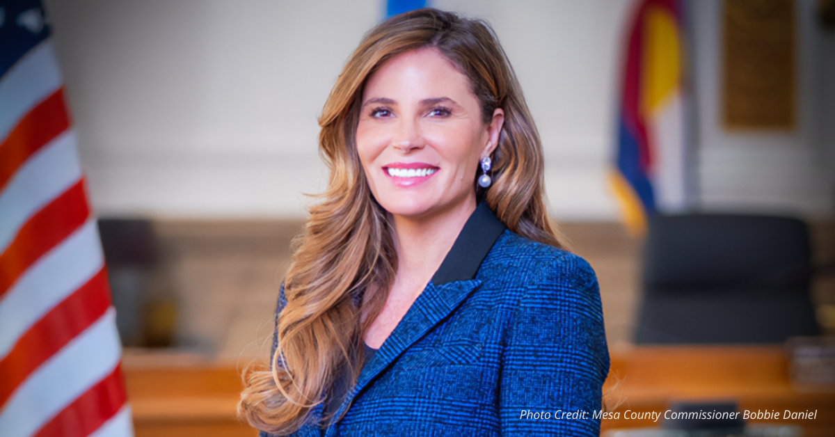 Smiling woman in a blue blazer standing indoors with an American flag in the background