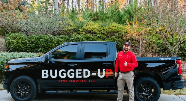 A technician in a red uniform gives a thumbs-up beside a black Bugged-Up Pest Services pickup truck outdoors.