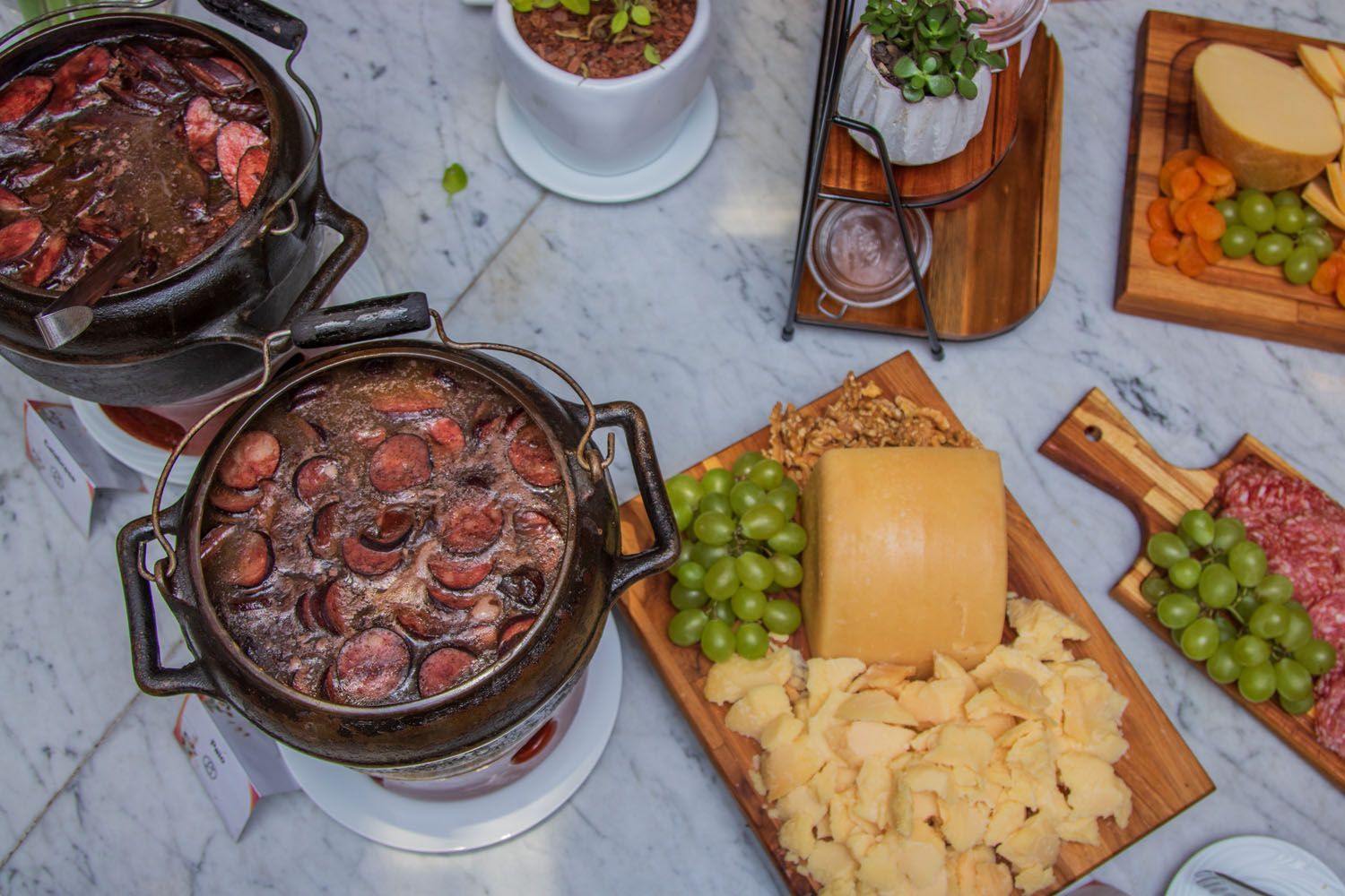 Feijoada com linguiça, queijo, uvas e salgadinhos em superfície de mármore branco.