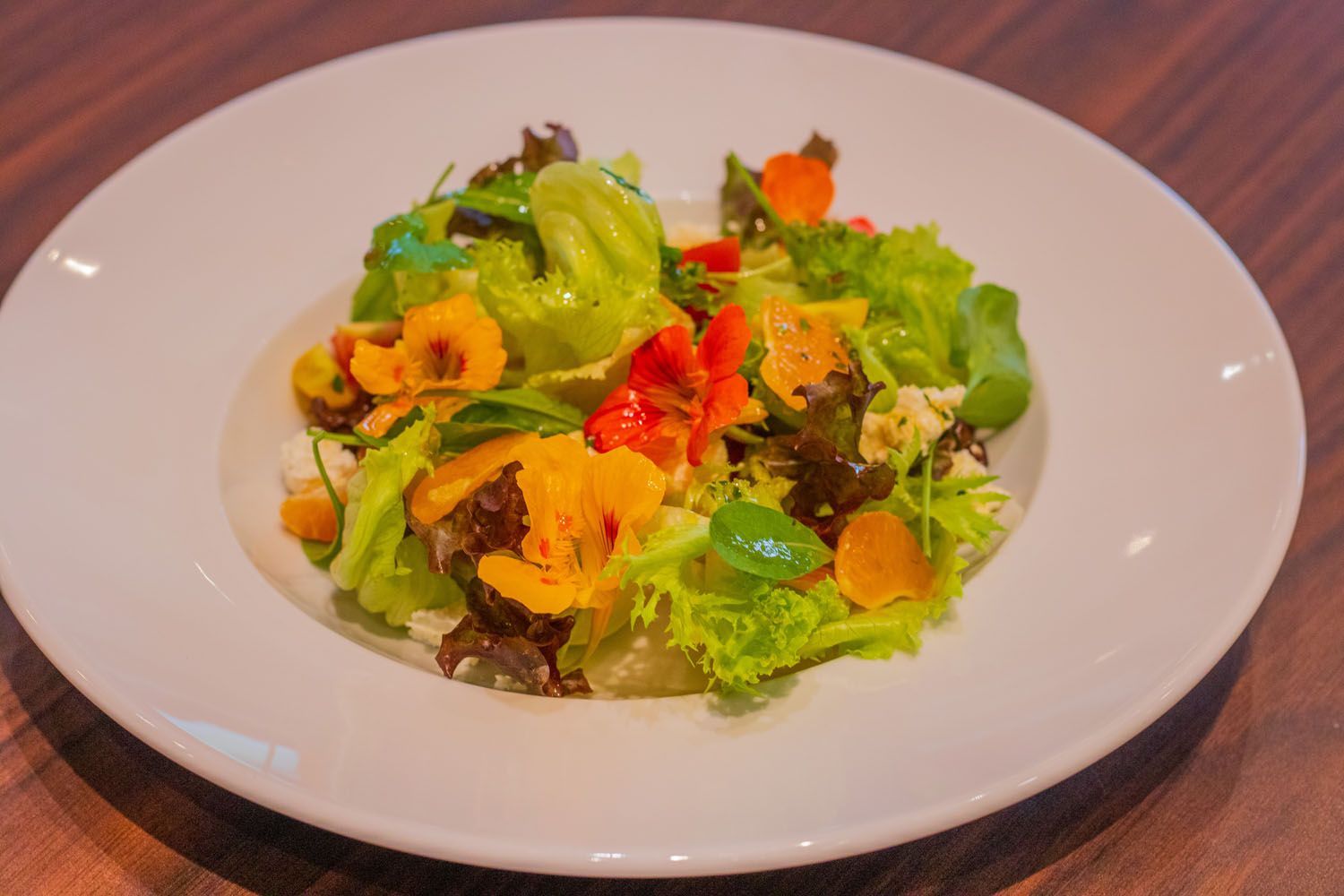 Salada em um prato branco com alface, flores, tomates e ervas. Mesa de madeira.