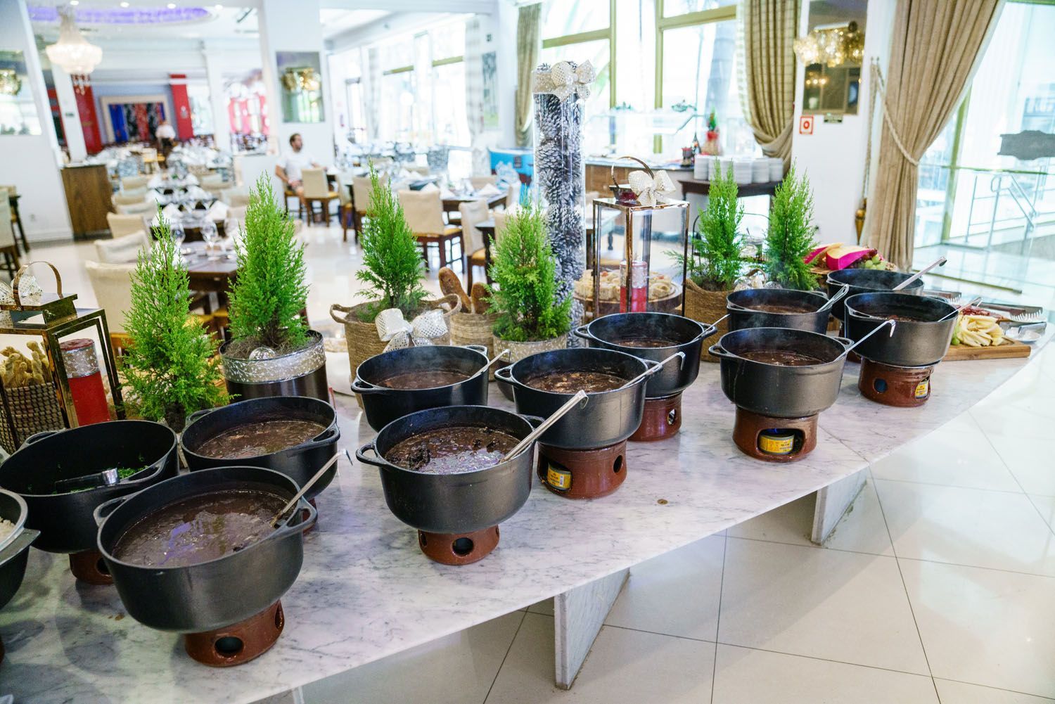 Buffet com várias panelas pretas de comida em suportes aquecidos, em uma área de jantar iluminada.