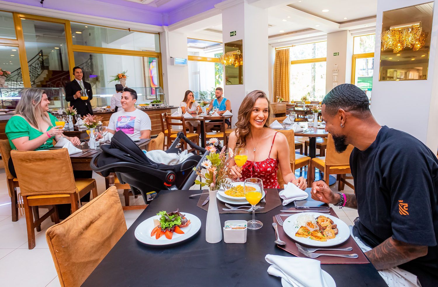 Pessoas jantando em um restaurante iluminado com mesas e cadeiras. Casal sorridente na entrada, carrinho de bebê por perto.