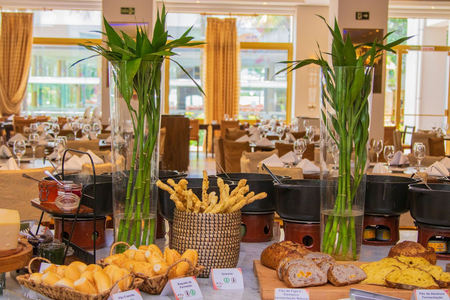 Buffet com pães, queijos e doces, decorado com plantas altas e verdes. Ambiente de restaurante, mesas ao fundo.