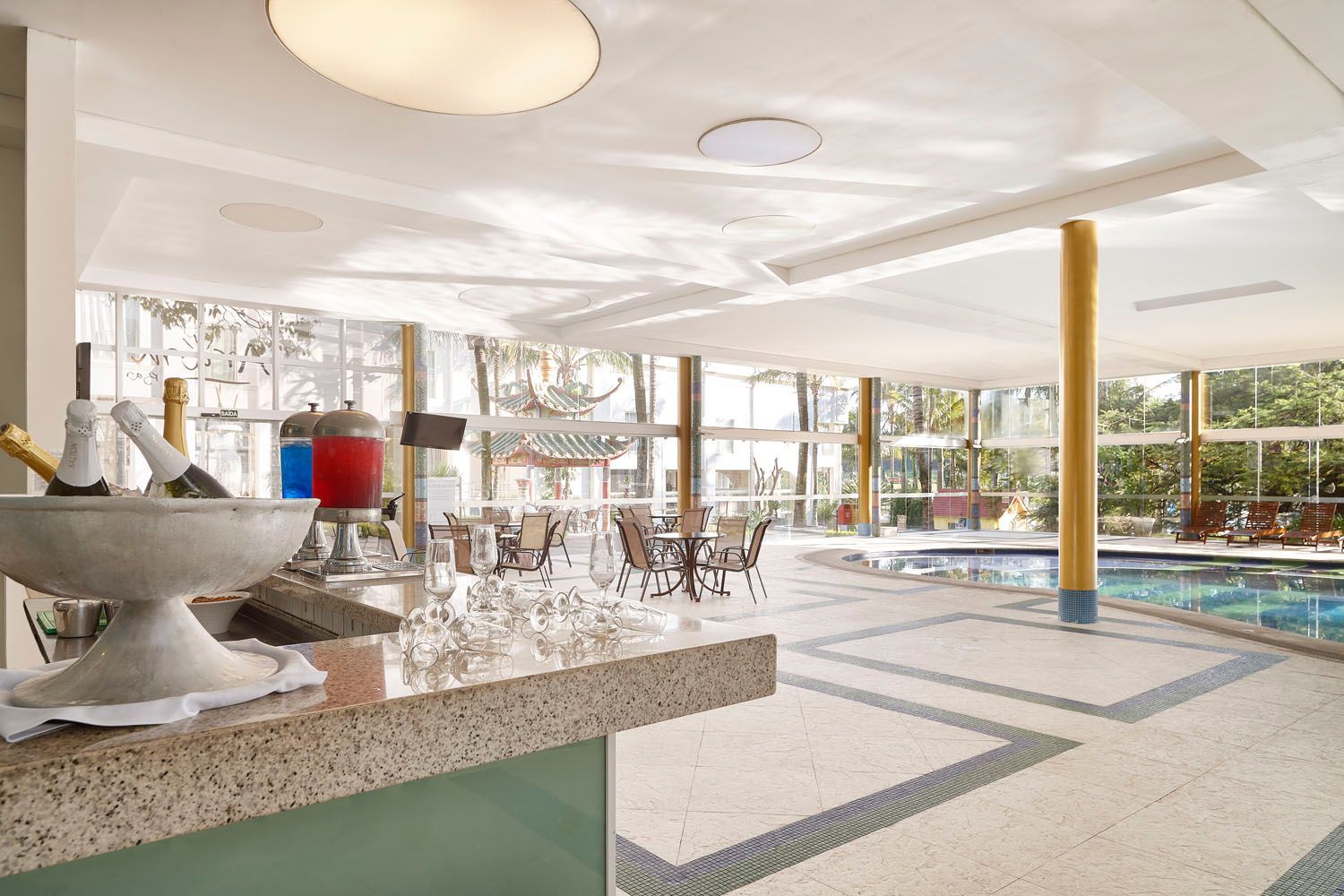 Área de piscina coberta com bar, balde de champanhe, mesas e piscina, com luz natural e vista para o exterior.