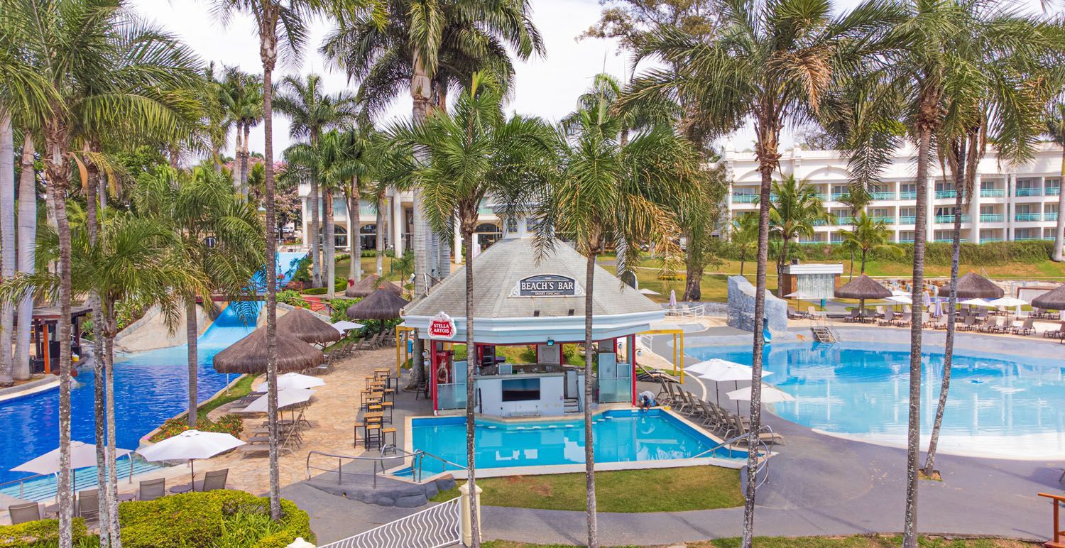 Bar à beira da piscina com piscinas azuis, palmeiras e edifícios brancos sob um céu brilhante.