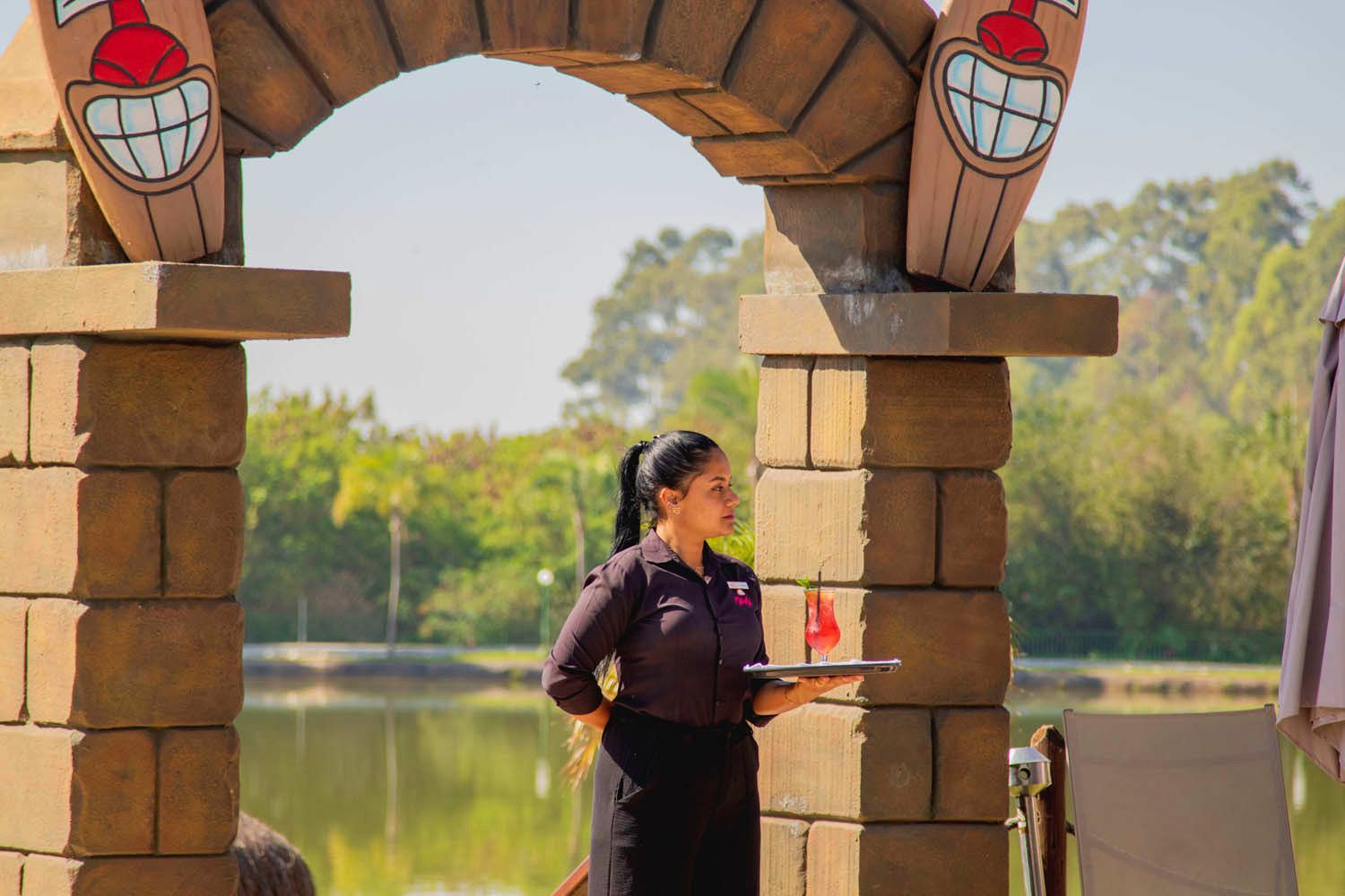 Garçonete segurando uma bandeja em pé sob um arco com tema tiki perto de um lago.