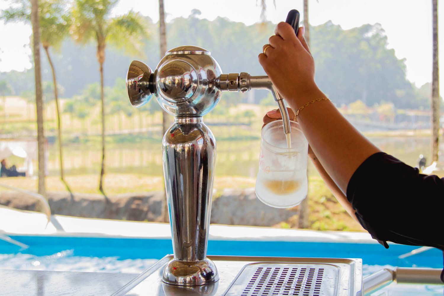 Barman servindo cerveja de uma torneira cromada, ambiente externo com vista para a piscina e o lago.