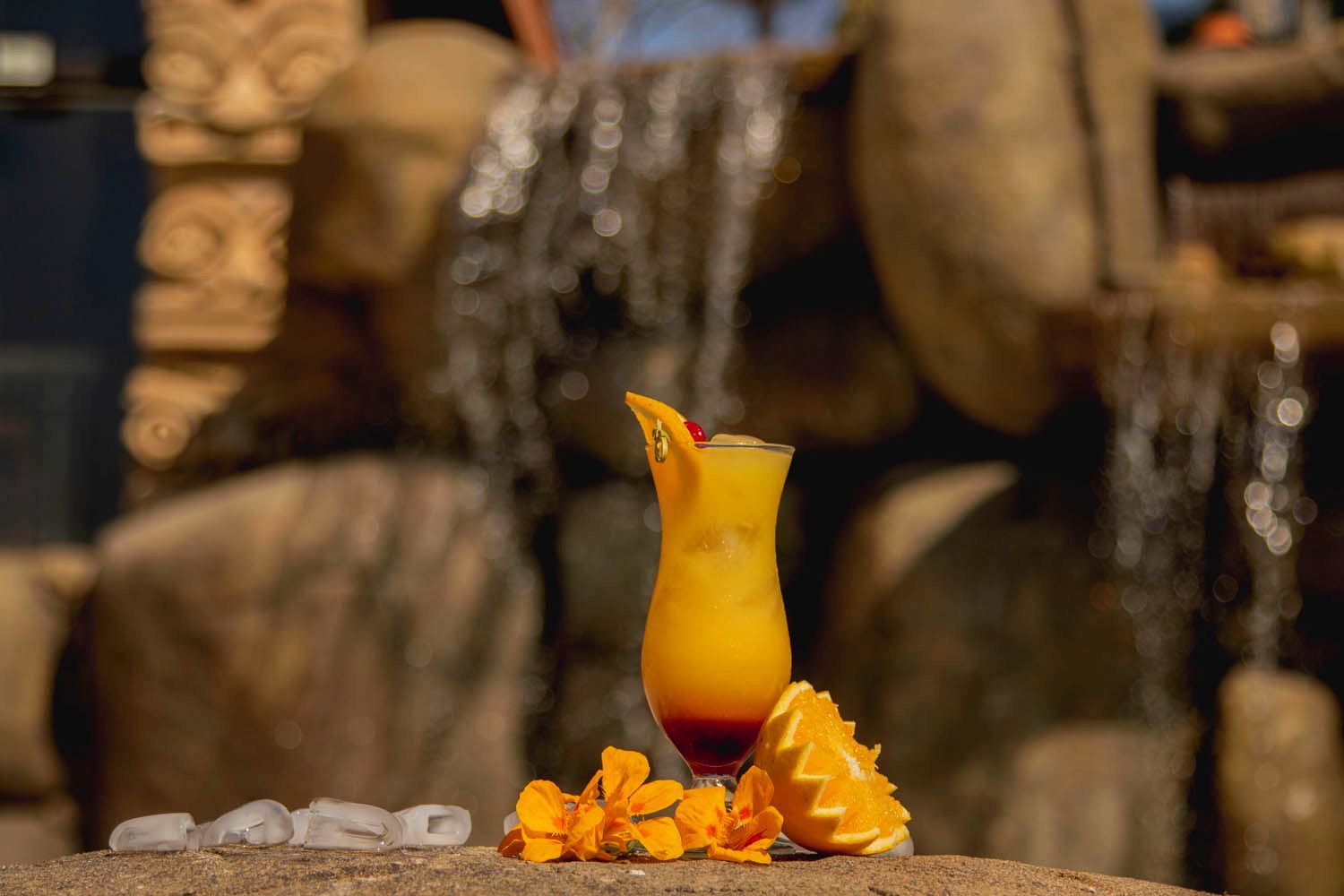 Coquetel tropical com fatia de laranja, cereja e flores sobre uma pedra, com cachoeira ao fundo.