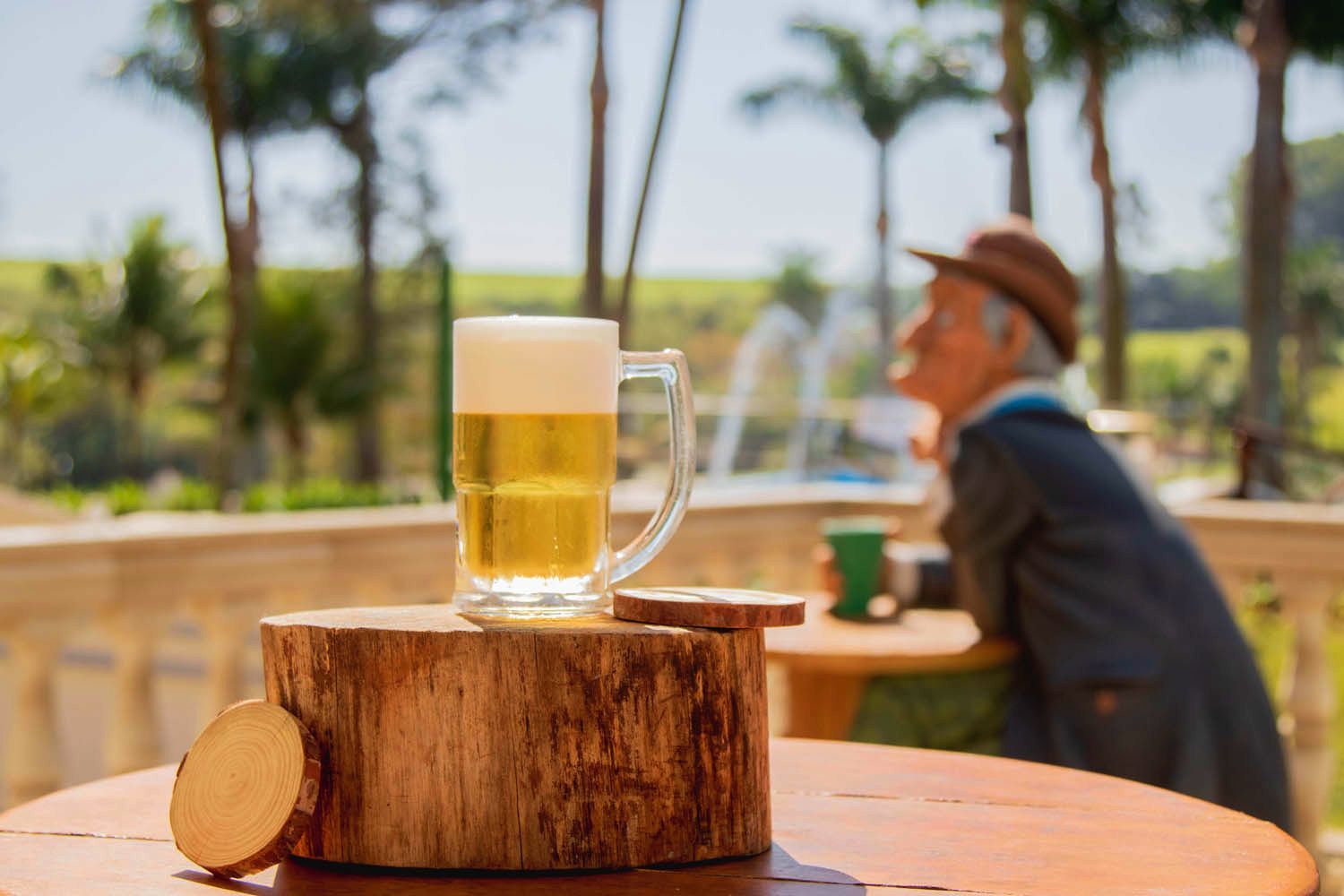Uma caneca de cerveja sobre um bloco de madeira, com um homem de chapéu desfocado ao fundo. Ambiente externo.