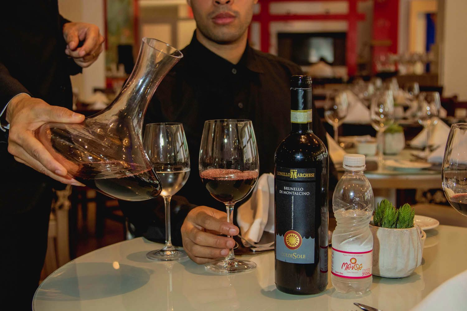 Um garçom servindo vinho tinto em uma taça, com uma pessoa sentada à mesa de um restaurante.