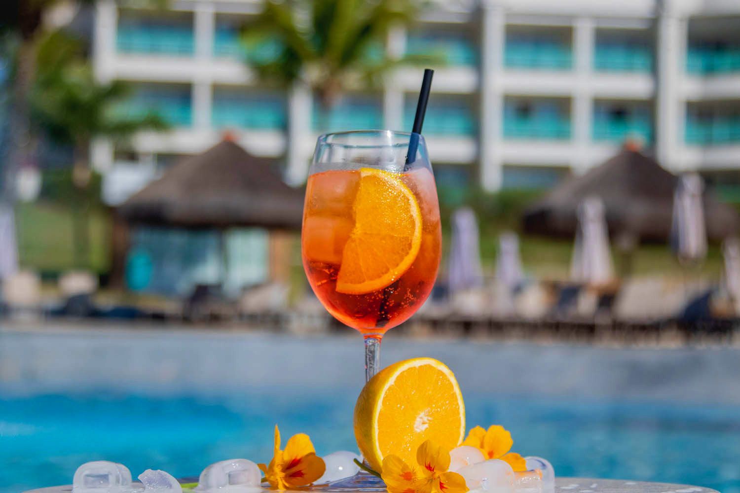 Coquetel com fatia de laranja e canudo à beira da piscina. Prédios do resort ao fundo.