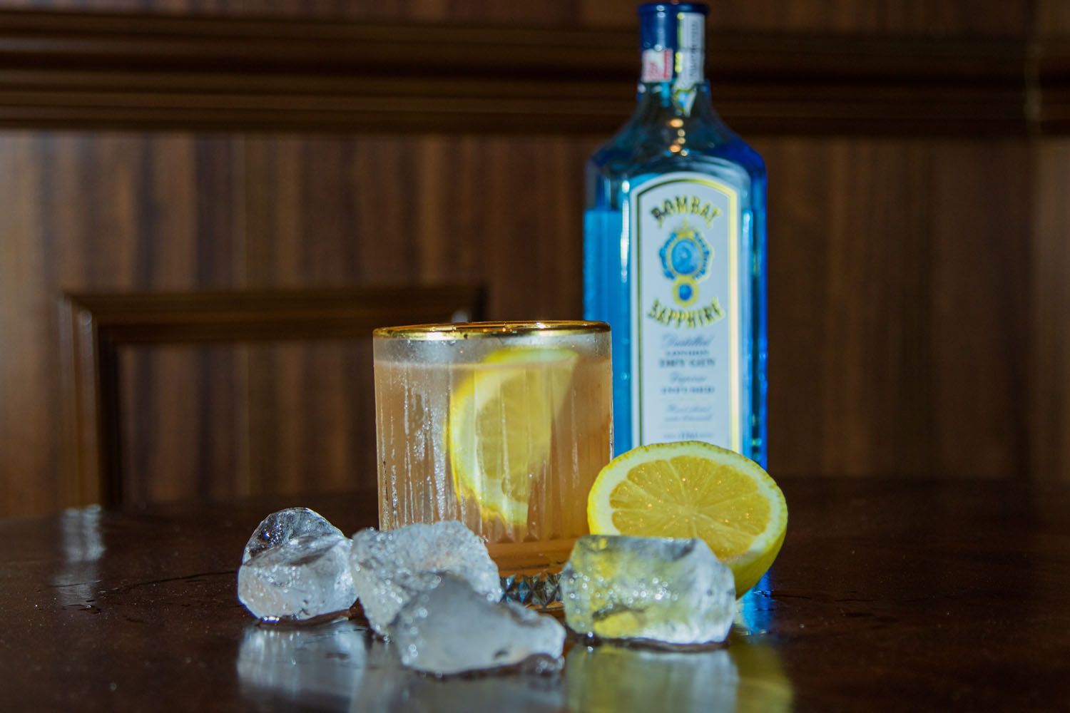 Um coquetel com fatia de limão, cubos de gelo e uma garrafa de gim Bombay Sapphire sobre uma superfície de madeira escura.