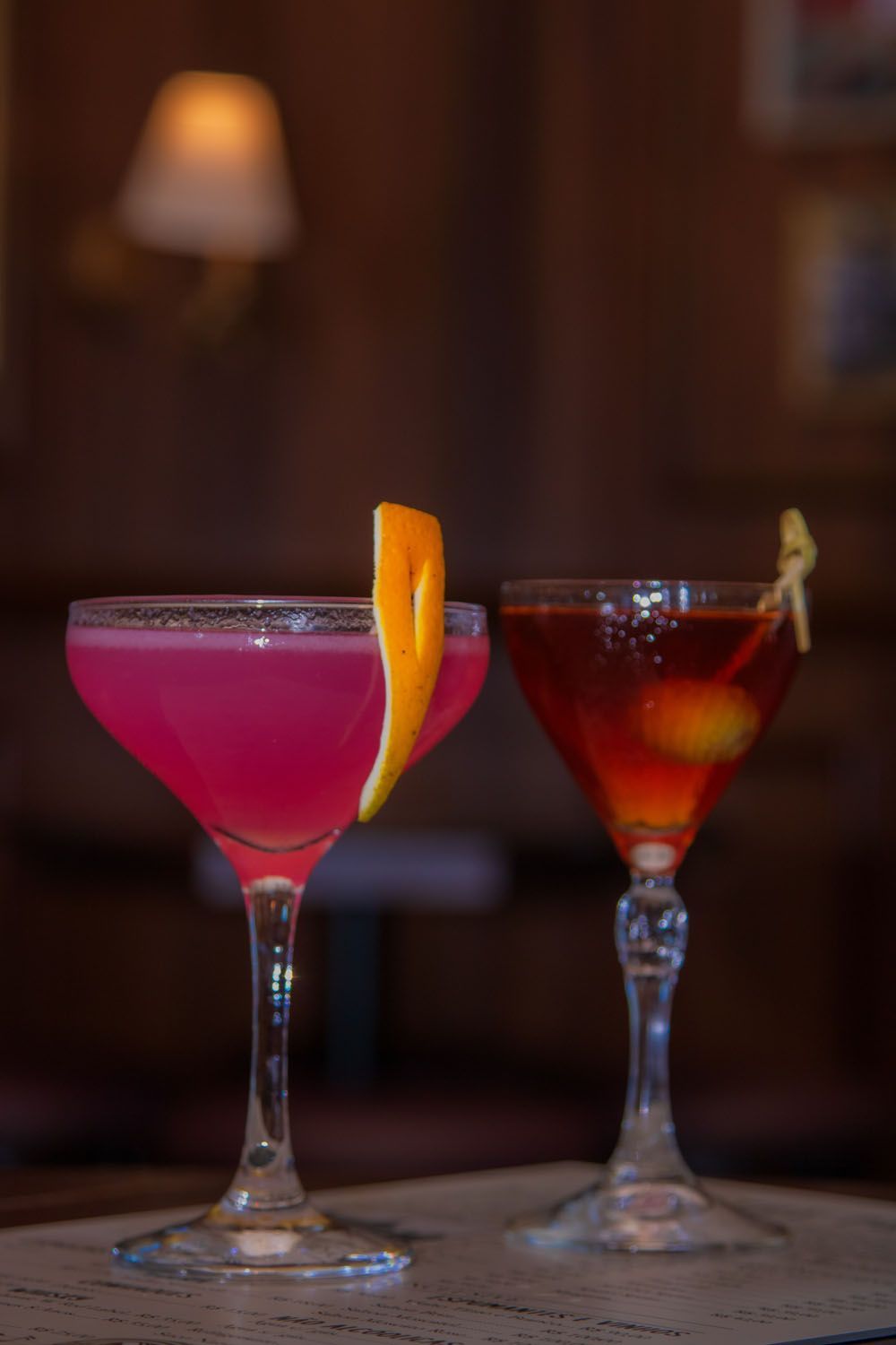 Dois coquetéis, um rosa com casca de laranja e outro tinto com azeitonas, em um bar.