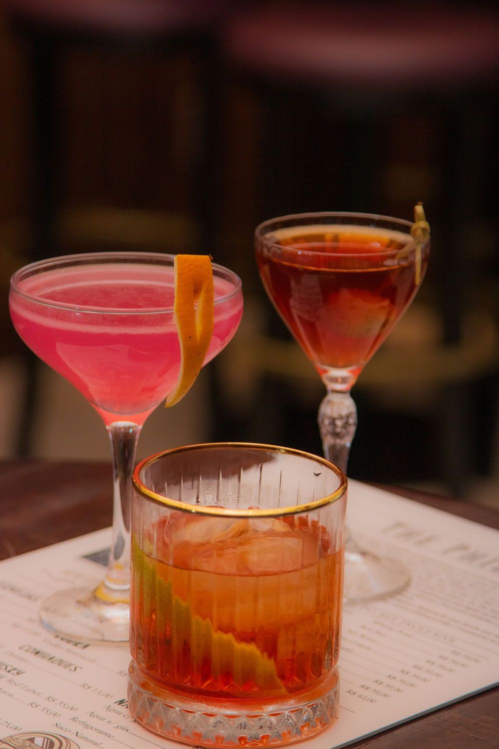 Três coquetéis coloridos sobre uma mesa: rosa, âmbar e laranja, decorados com frutas cítricas em um ambiente de bar com pouca luz.