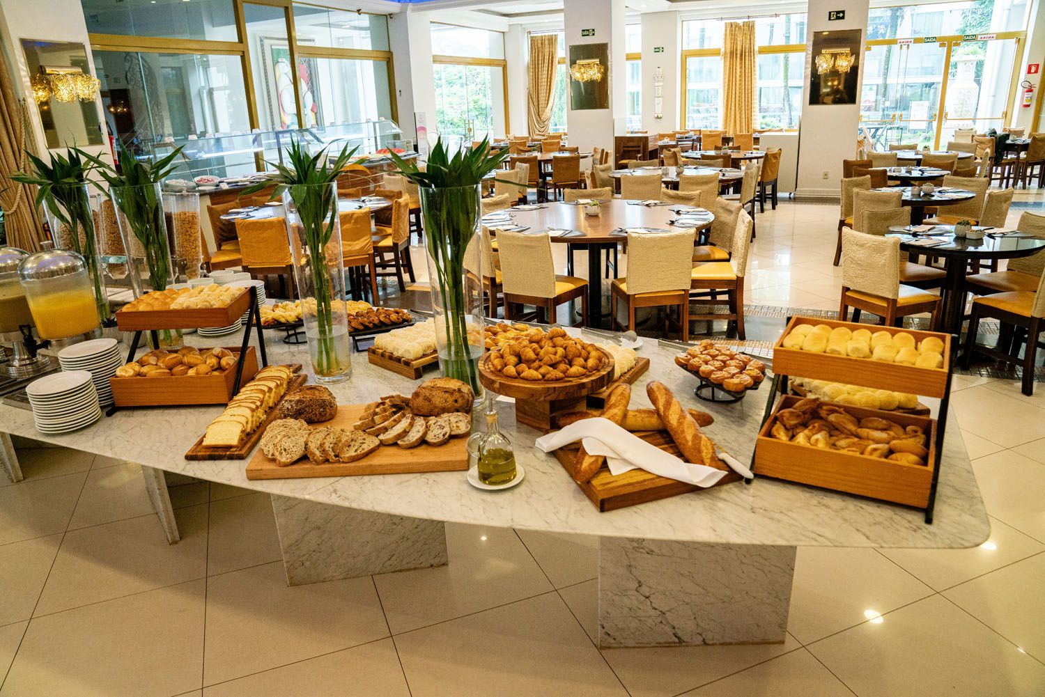Buffet com diversos pães e doces, ambiente de restaurante com mesas e cadeiras.