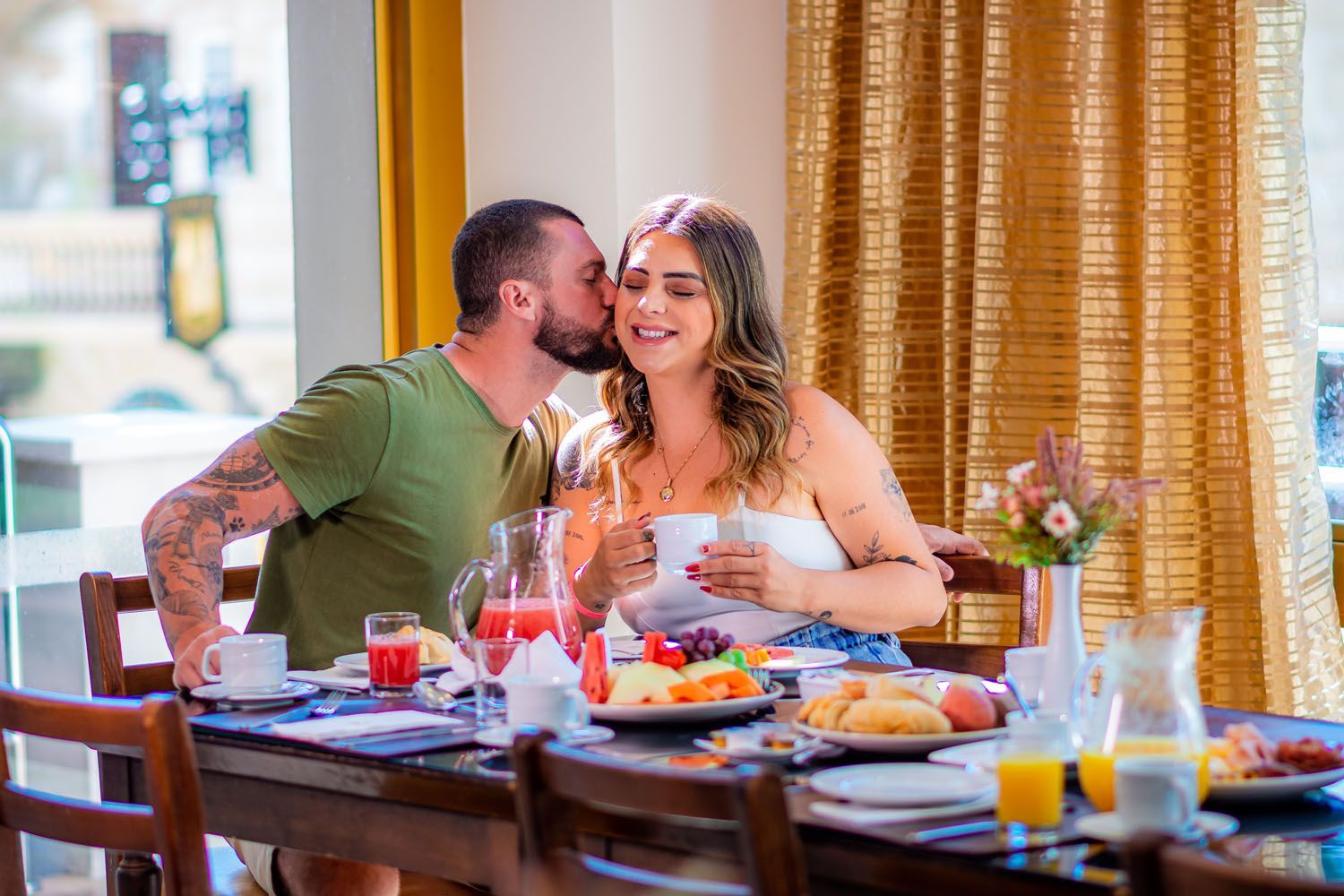 Homem beija a bochecha de mulher na mesa de café da manhã, com comida e bebida em um ambiente iluminado.
