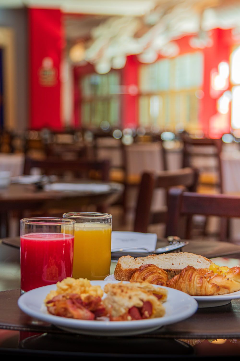 Café da manhã com suco, torradas, doces e mesas de restaurante ao fundo.