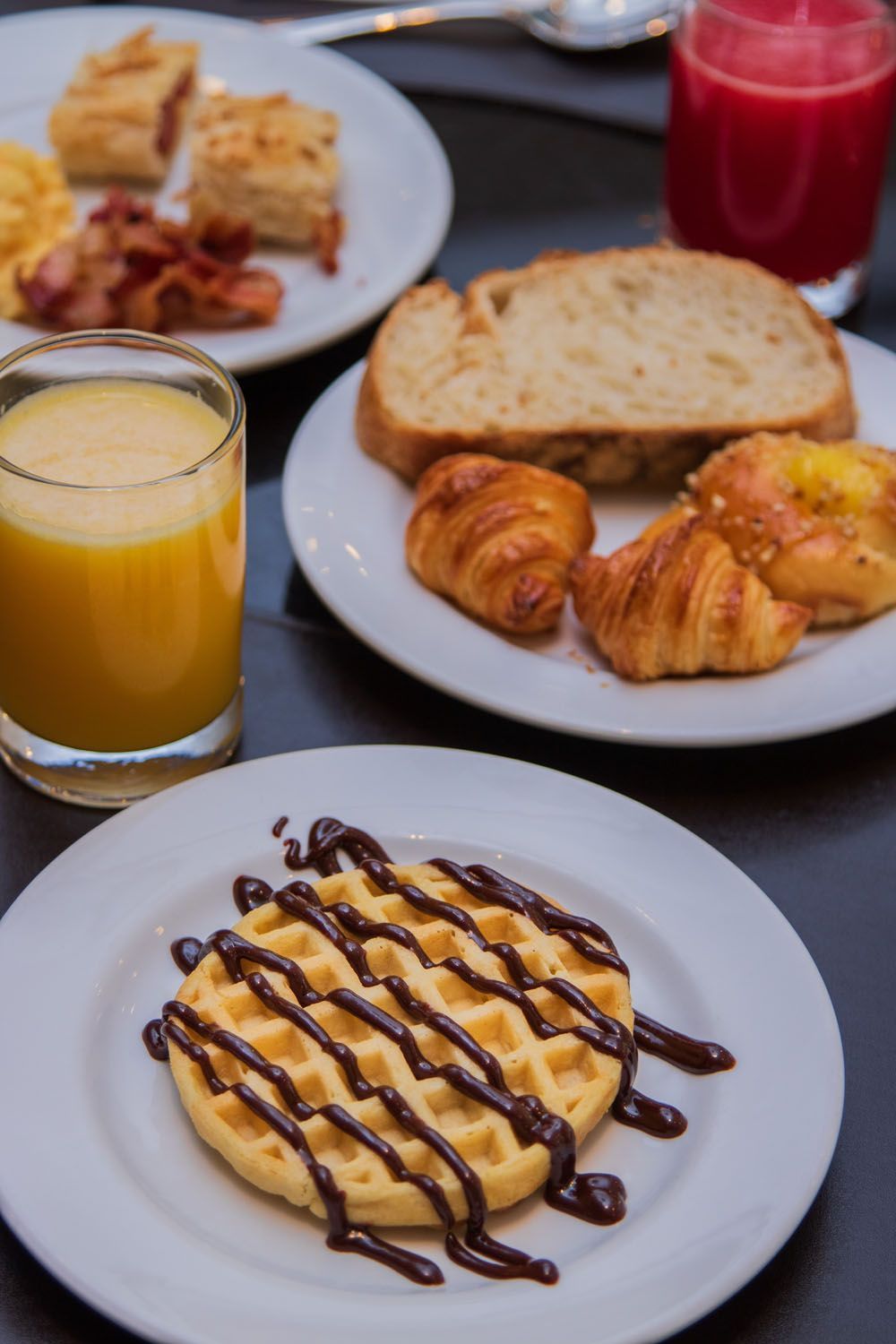 Café da manhã com waffle, doces, torradas, bacon, ovos e suco.