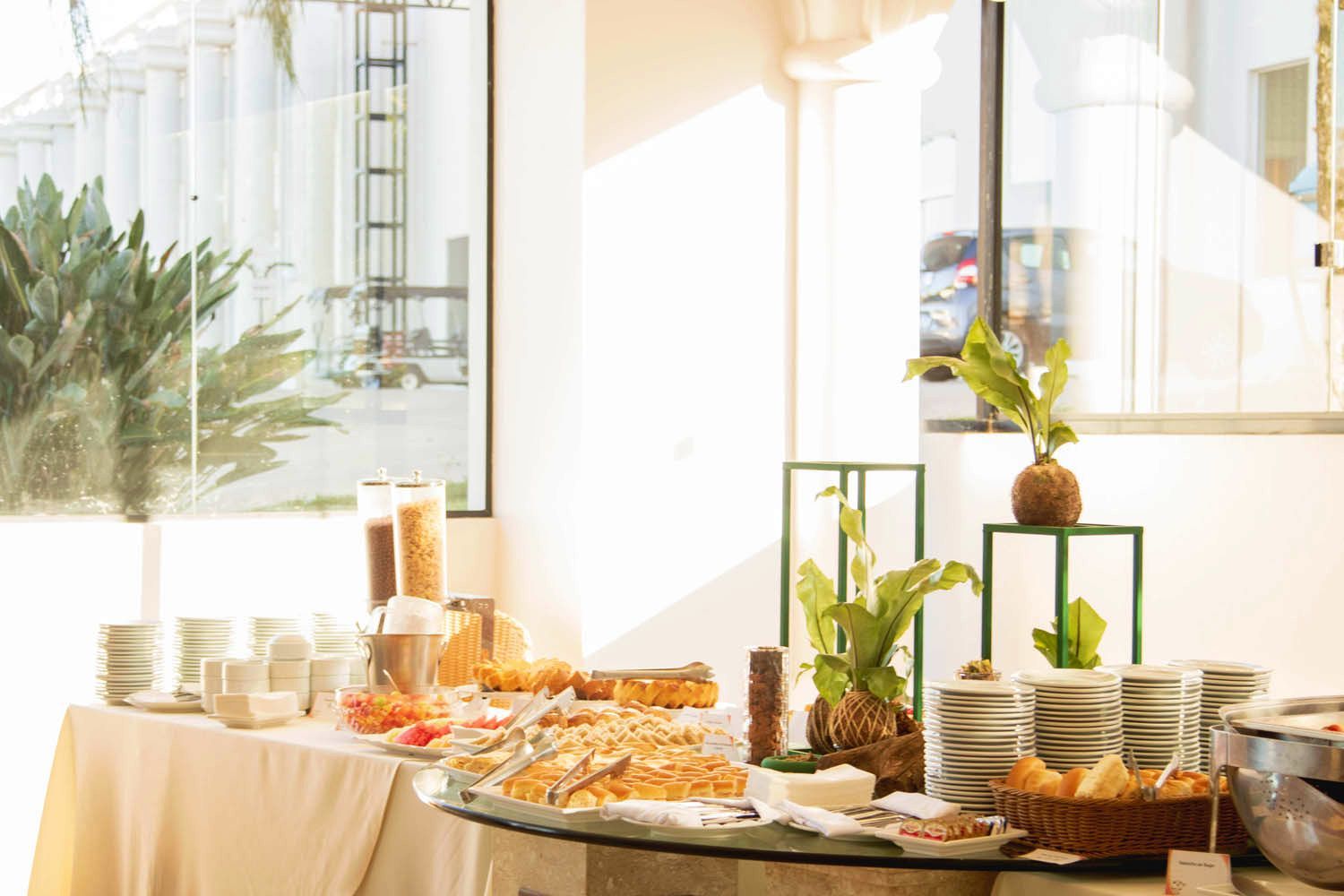 Buffet de café da manhã em um ambiente bem iluminado, com comida, pratos e plantas na mesa. Grandes janelas com vista para uma área industrial.