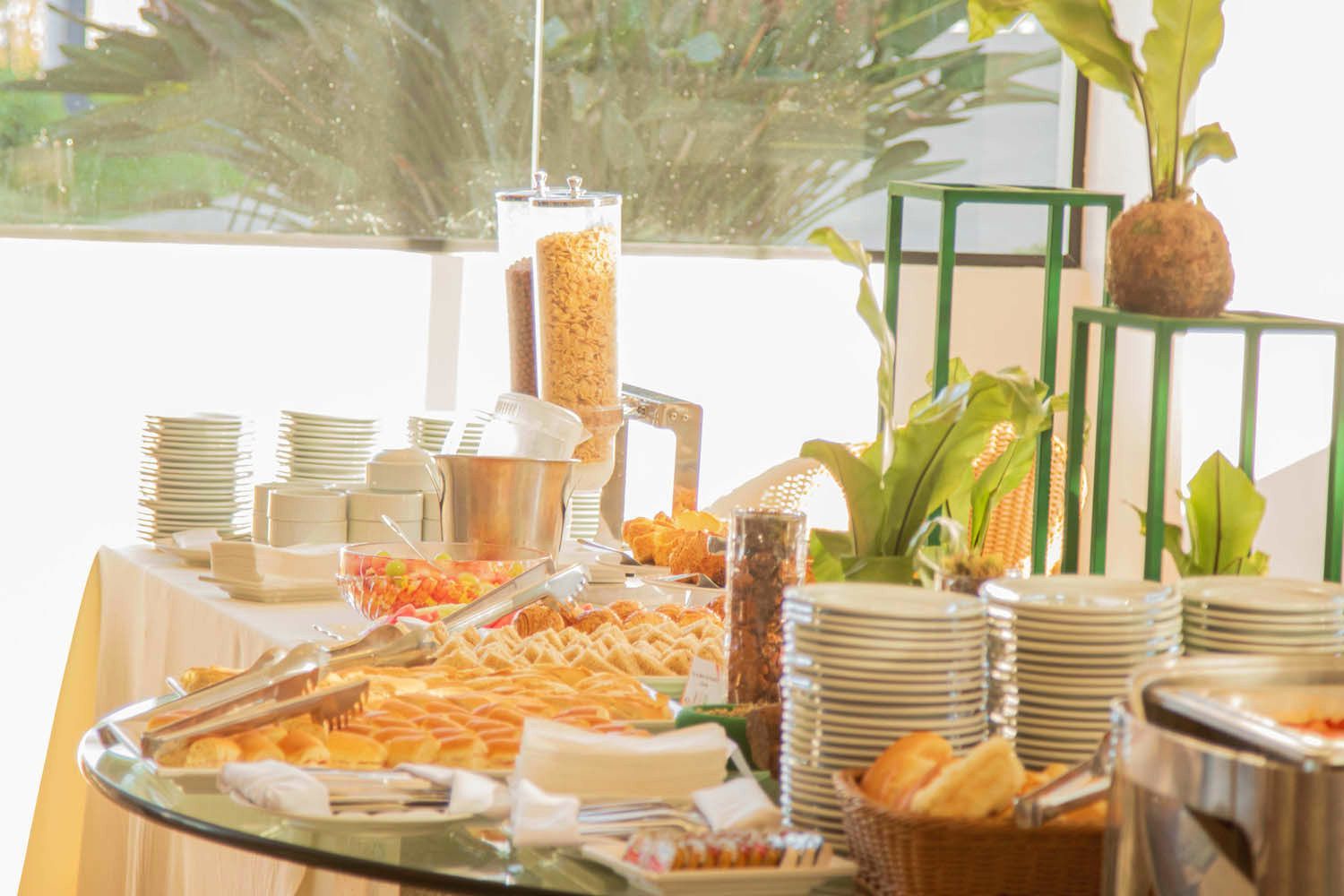 Buffet de café da manhã com diversos doces, cereais e frutas. Inclui pratos brancos e toalha de mesa bege.