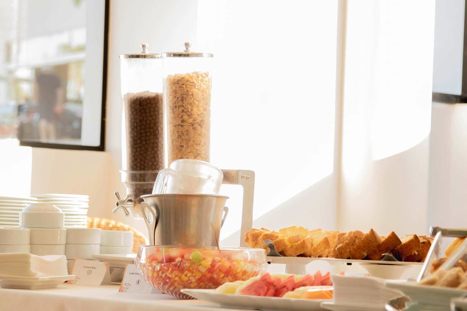 Buffet de café da manhã com dispensadores de cereais, frutas e doces em uma toalha de mesa branca.