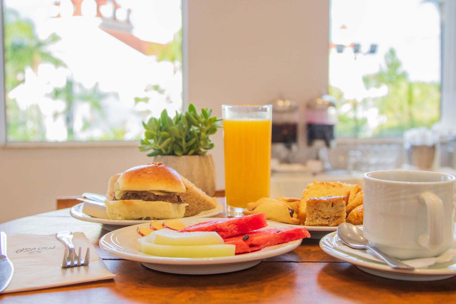 Mesa de café da manhã: hambúrguer, fatias de frutas, suco de laranja, doces, café, com vista para a janela.