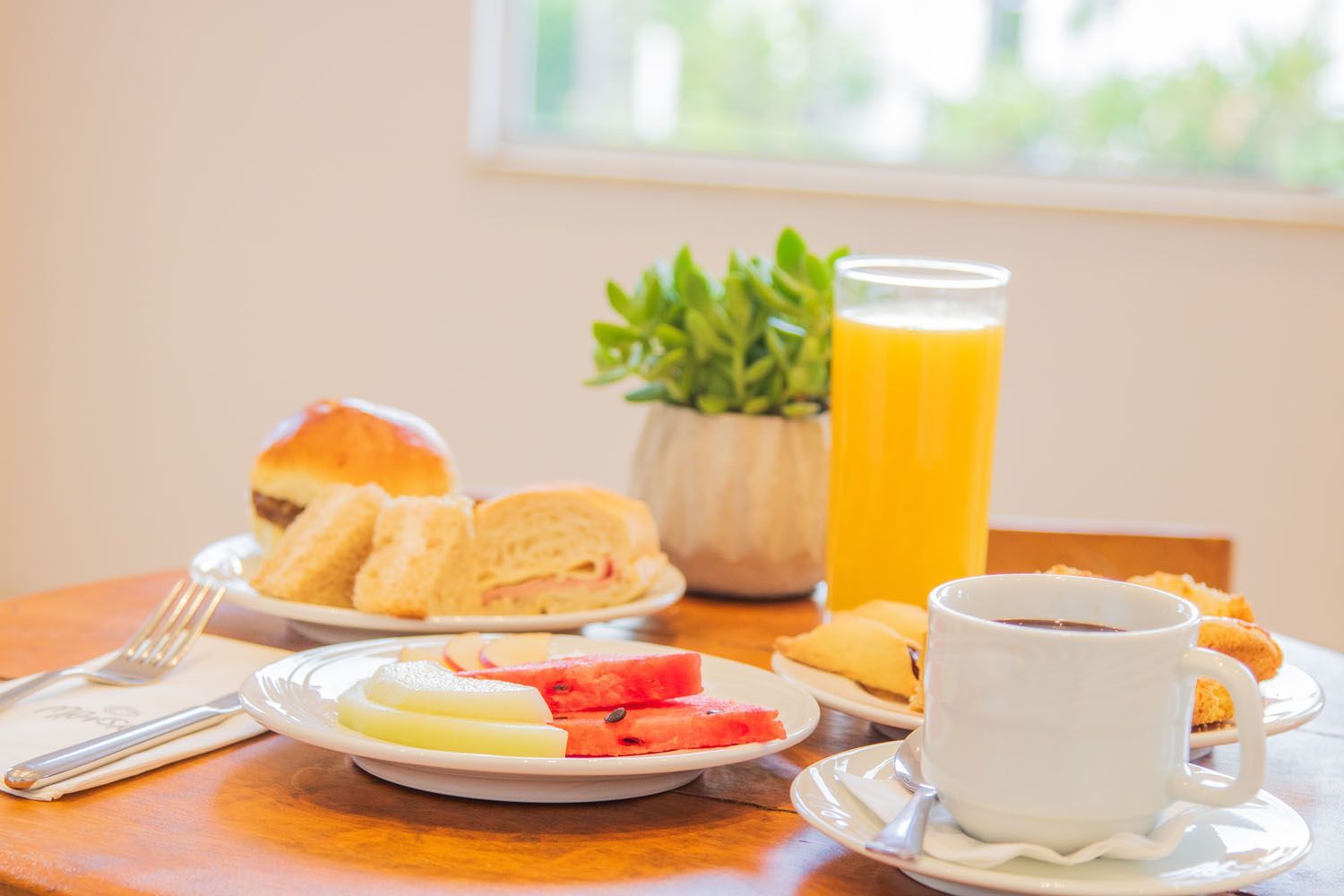 Café da manhã com sanduíches, frutas, café e suco de laranja em uma mesa de madeira.
